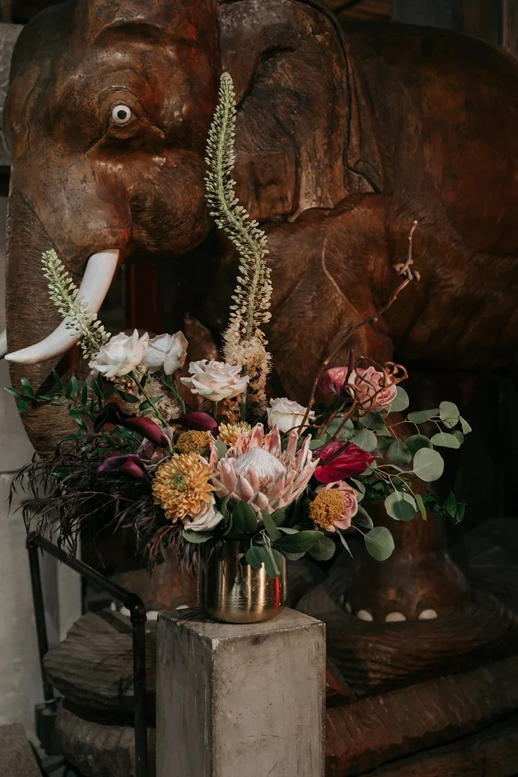 A bronze-colored elephant sculpture with white tusks and large ears, behind a floral arrangement with various flowers and greenery, placed on a concrete block.