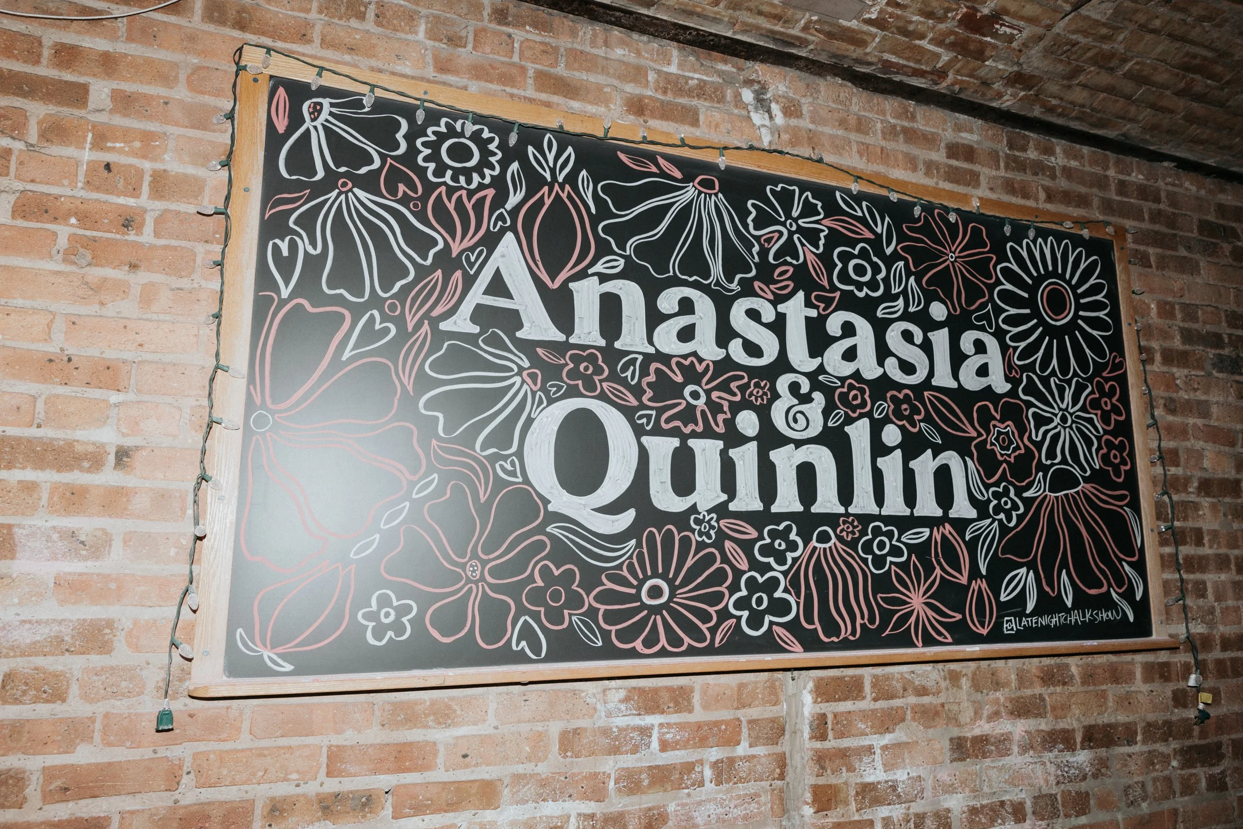 Chalkboard sign with floral illustrations and the text 'Anastasia & Quinton' hanging on a brick wall, illuminated by string lights.