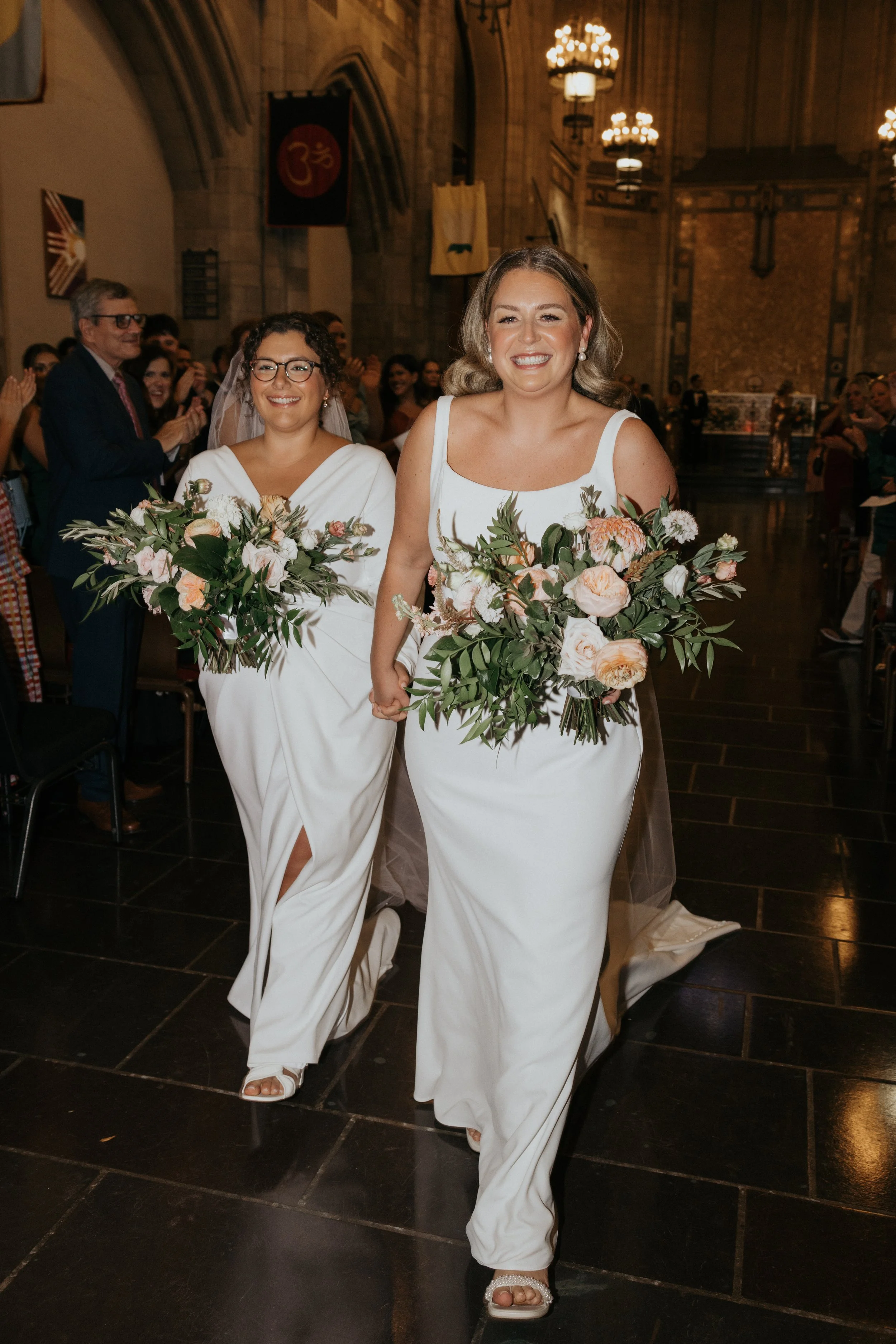 Two women in white dresses walking down the aisle at a wedding ceremony, holding hands and smiling, with guests clapping and taking photos in a church with high ceilings and chandeliers.