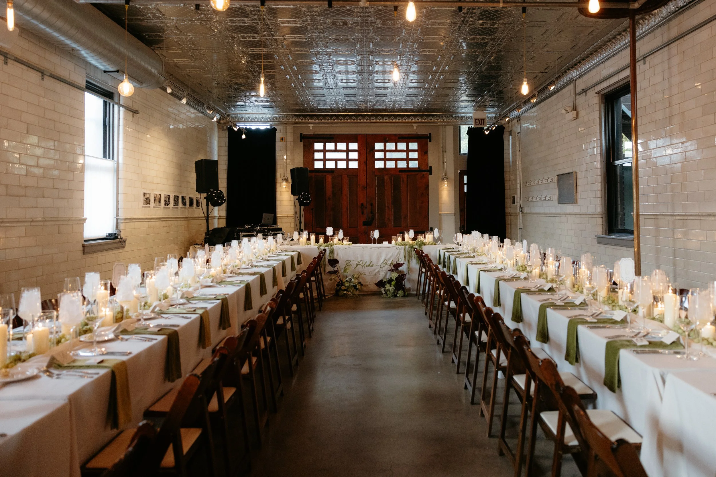 Wedding reception venue with long tables decorated with candles, glassware, and green and white table settings, set for a celebration in a rustic indoor space with exposed brick walls and wooden doors.