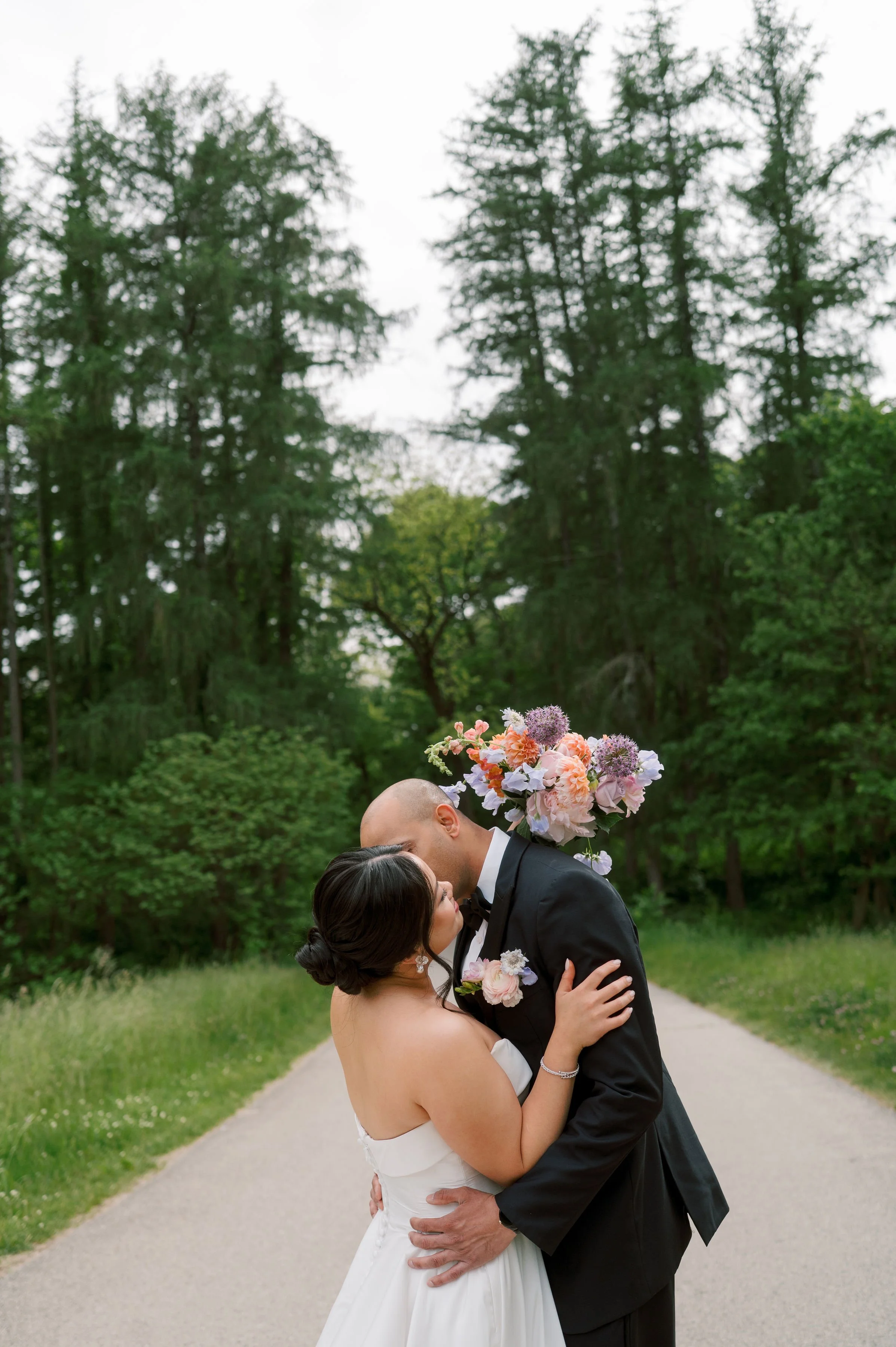 A bride and groom sharing a kiss outdoors on a paved path, with green trees and grass in the background, the bride is wearing a white wedding gown and the groom is in a black tuxedo with a floral boutonniere.