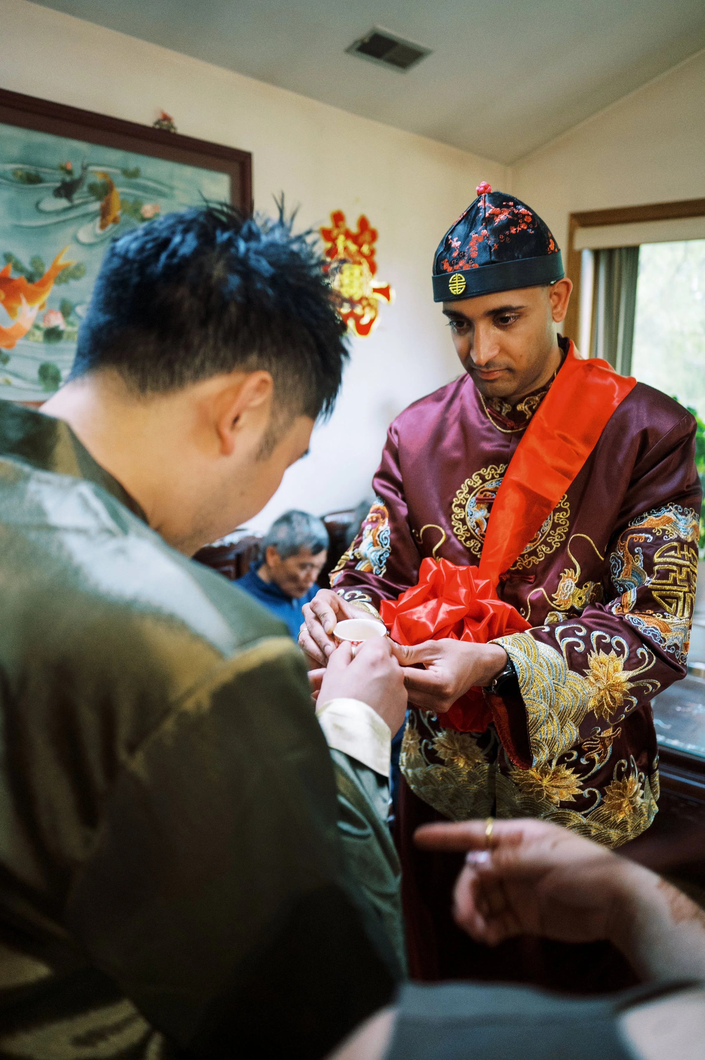 A man dressed in traditional Asian attire with intricate golden embroidery, wearing a black and red floral hat, holding a small cup with a red ribbon, participating in a cultural ceremony or celebration indoors.