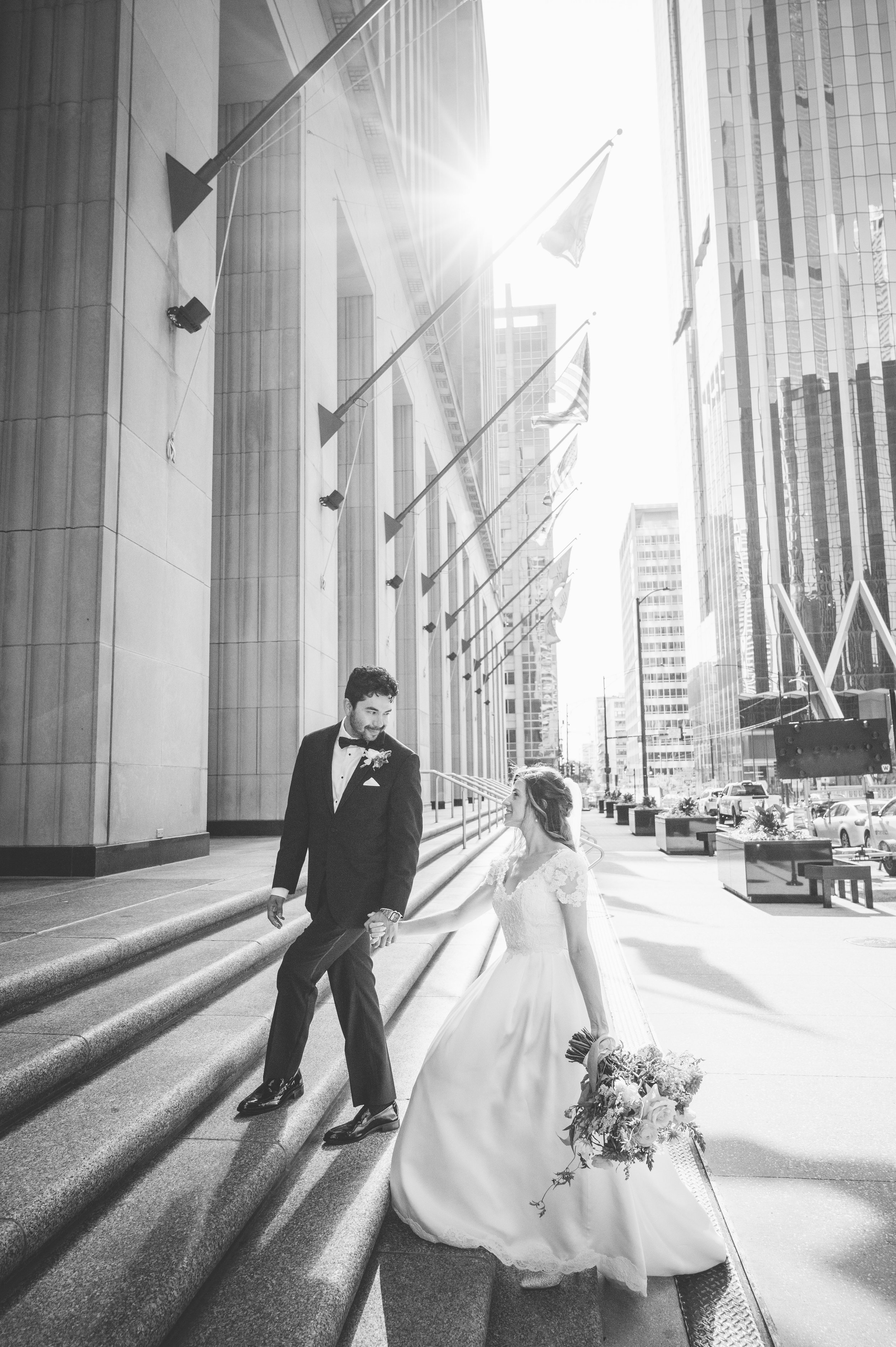 A bride and groom are holding hands and looking at each other outside a building with American flags and tall glass buildings in the background.