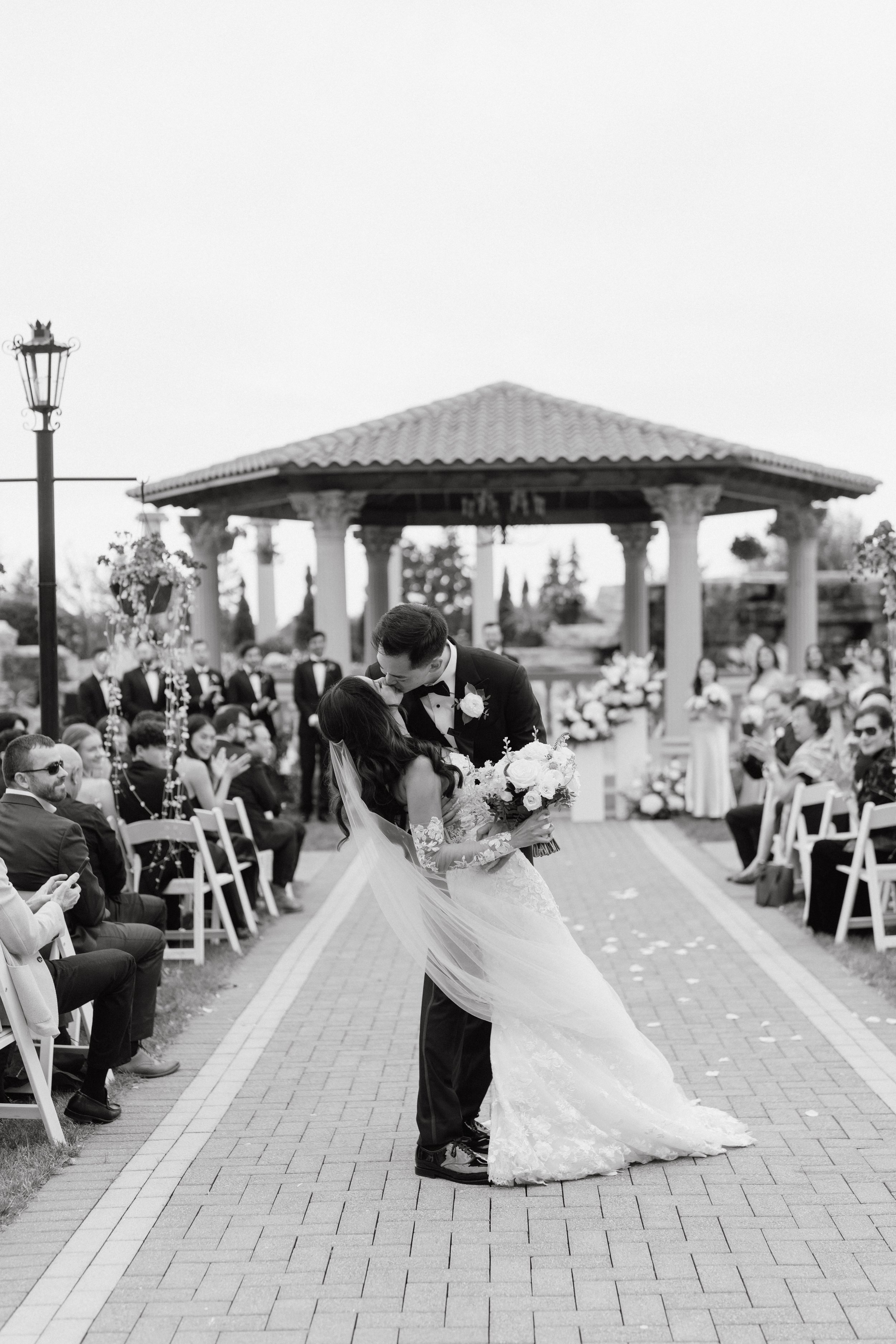 A bride and groom kiss at their outdoor wedding ceremony with guests seated and some standing, a gazebo in the background, and a lamppost on the left, captured in black and white.
