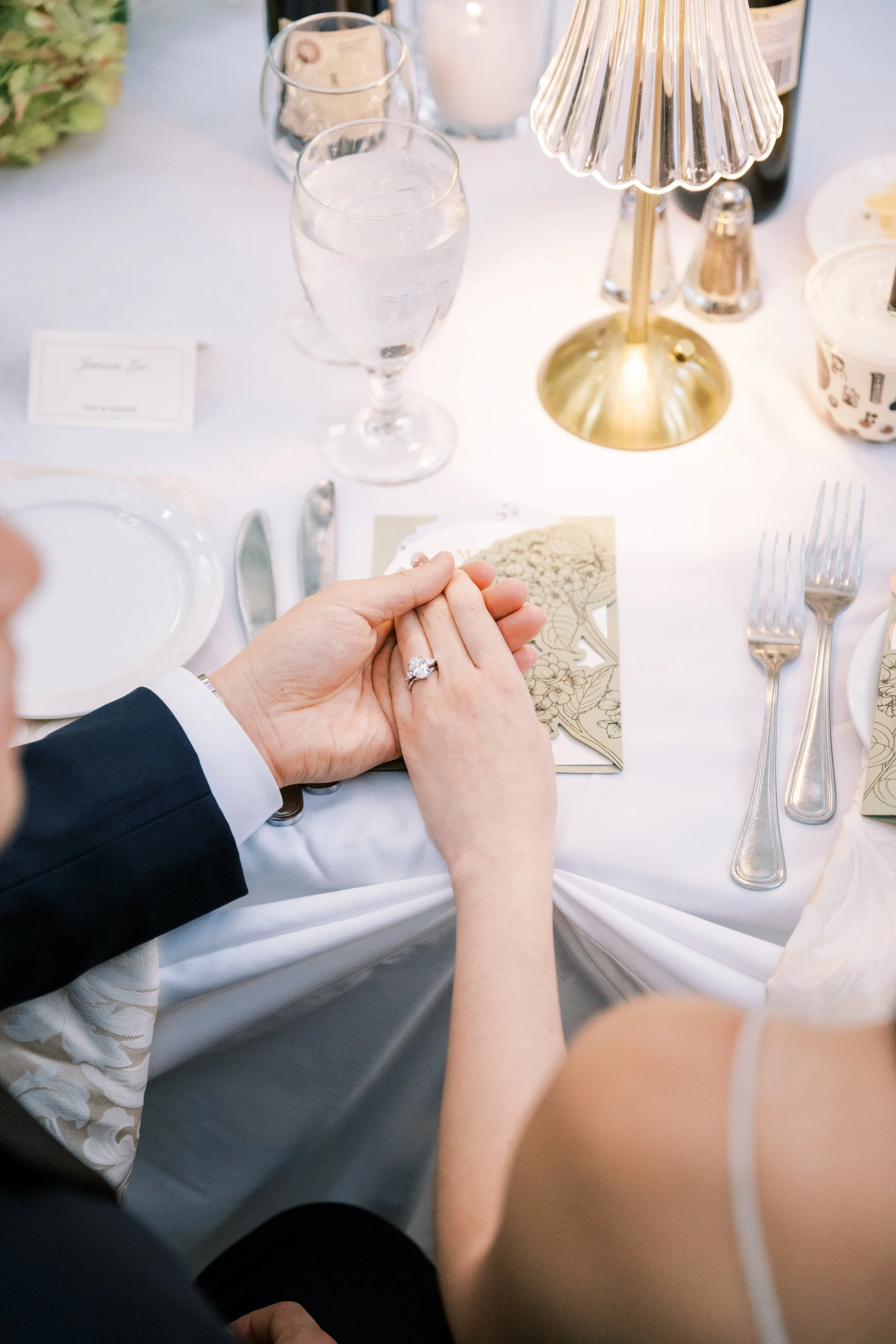 A wedding reception table with two drinks, a gold lamp, a guest wearing a dark suit, and a bride with a diamond ring holding hands with her groom.