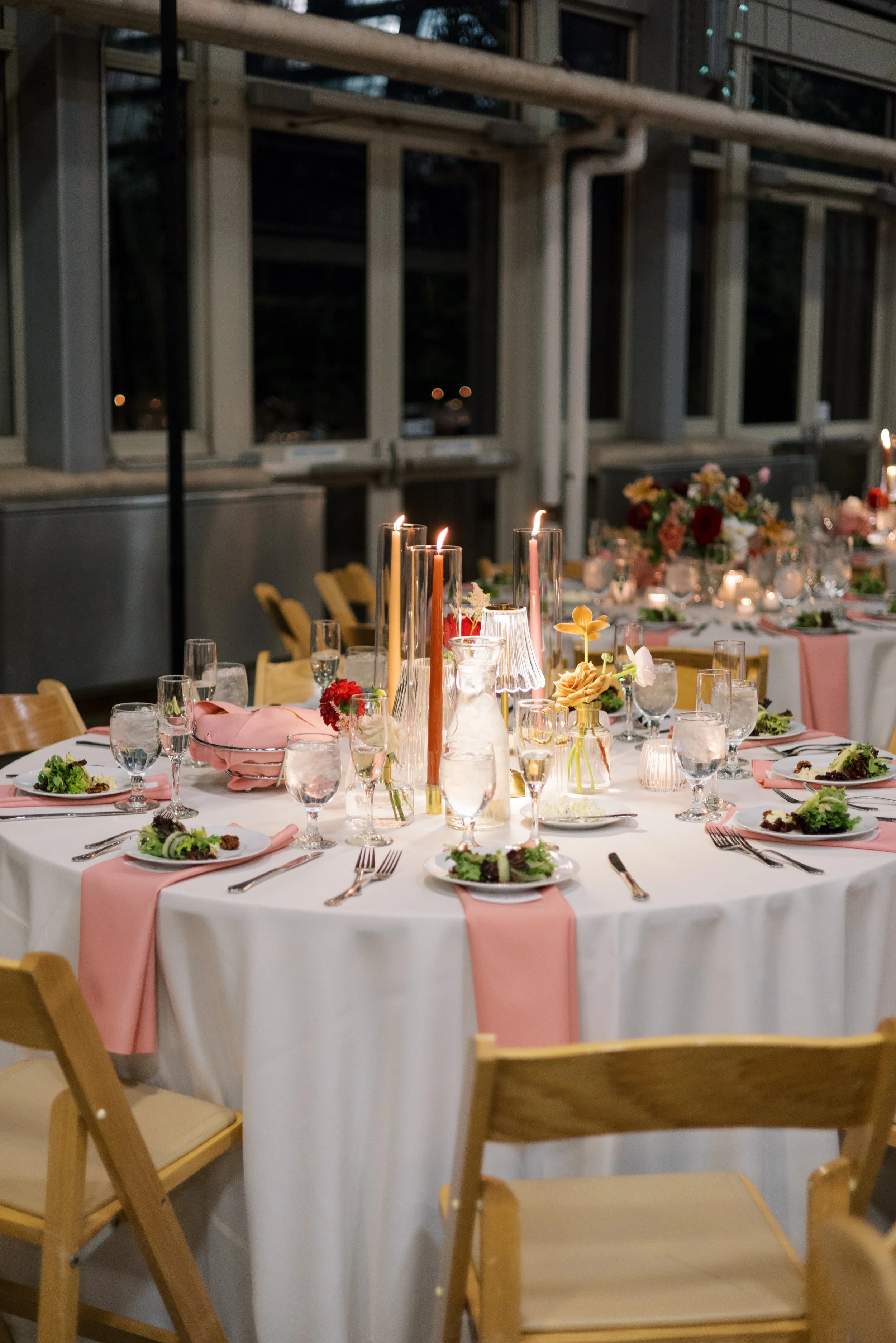 A decorated round dining table set for a formal event with pink tablecloths, floral centerpieces, and tall candles inside glass holders, surrounded by wooden chairs in a dimly lit room with large windows.