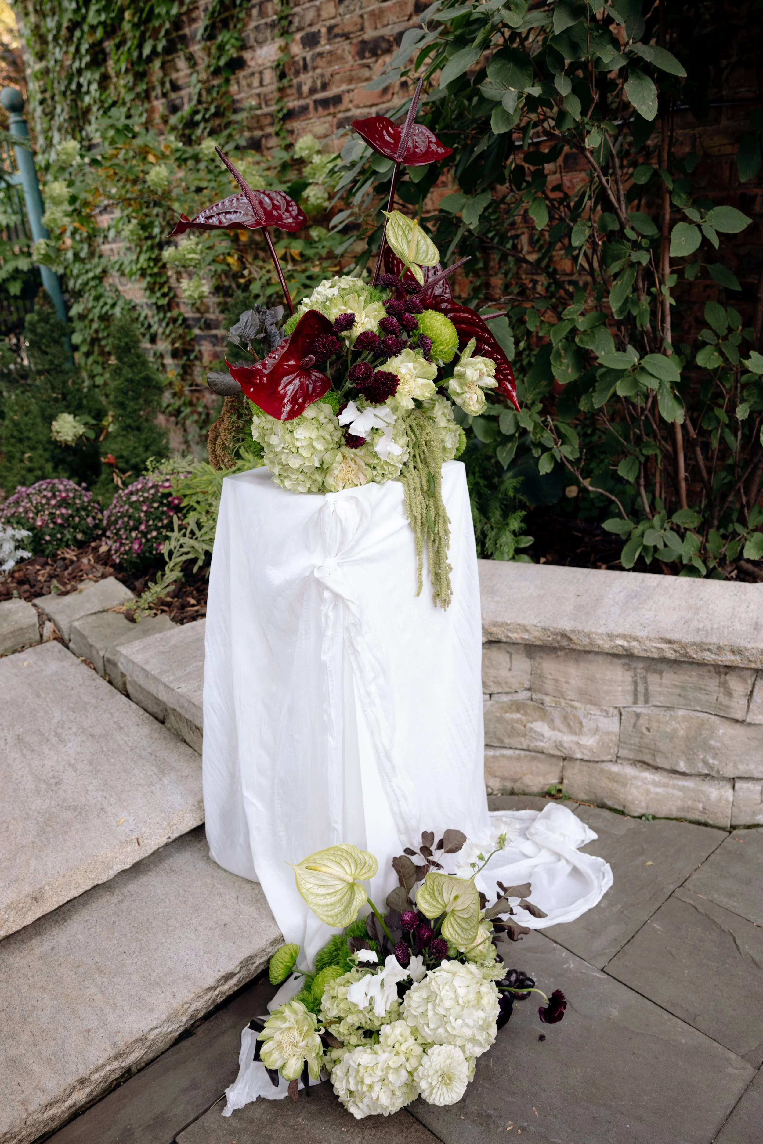 A floral arrangement on a white draped pedestal outdoors, featuring large white hydrangeas, dark red anthuriums, dark purple small flowers, and green foliage, with a brick wall and green plants in the background.
