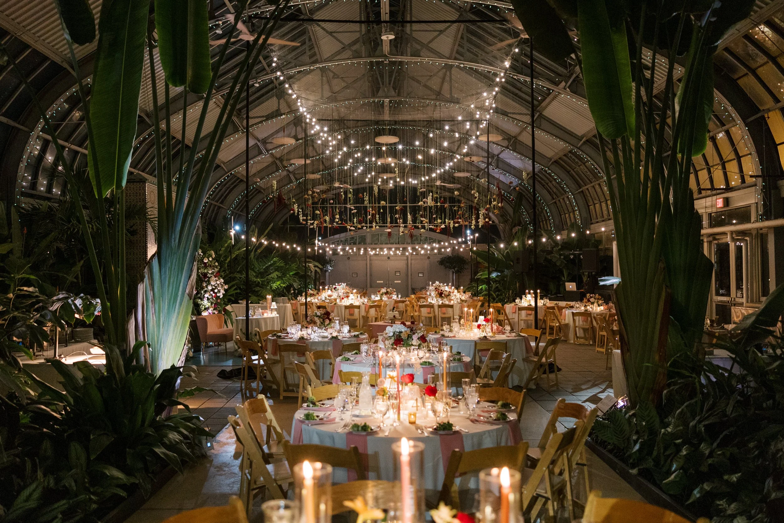 Indoor event space decorated for a celebration with string lights, hanging floral installations, and tables set for dining.