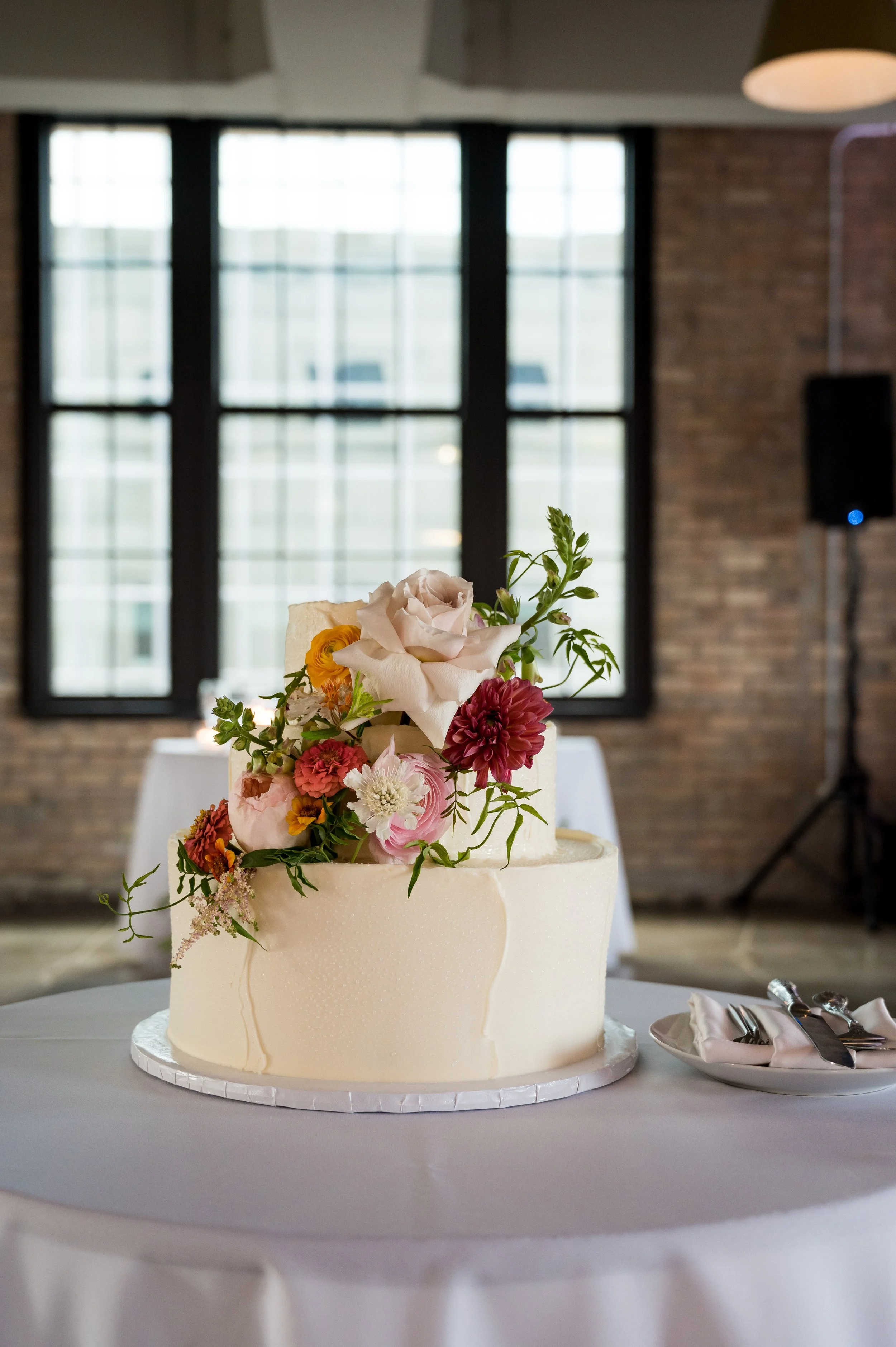 A white, two-tiered wedding cake decorated with flowers, including roses and dahlias, on a white tablecloth in a venue with large windows and exposed brick walls.