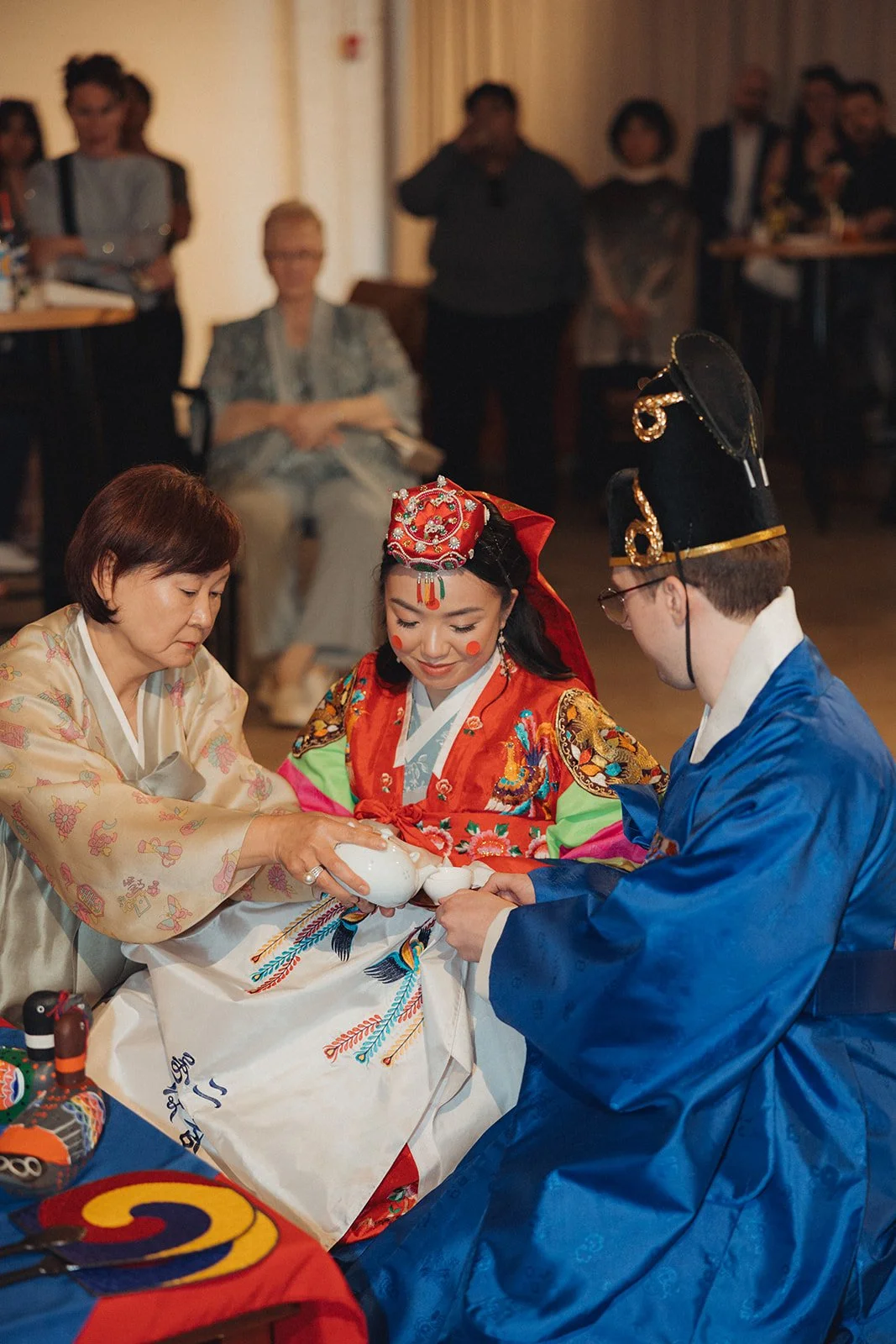 Three people in traditional Korean attire pouring tea during a ceremony, with several onlookers in the background.