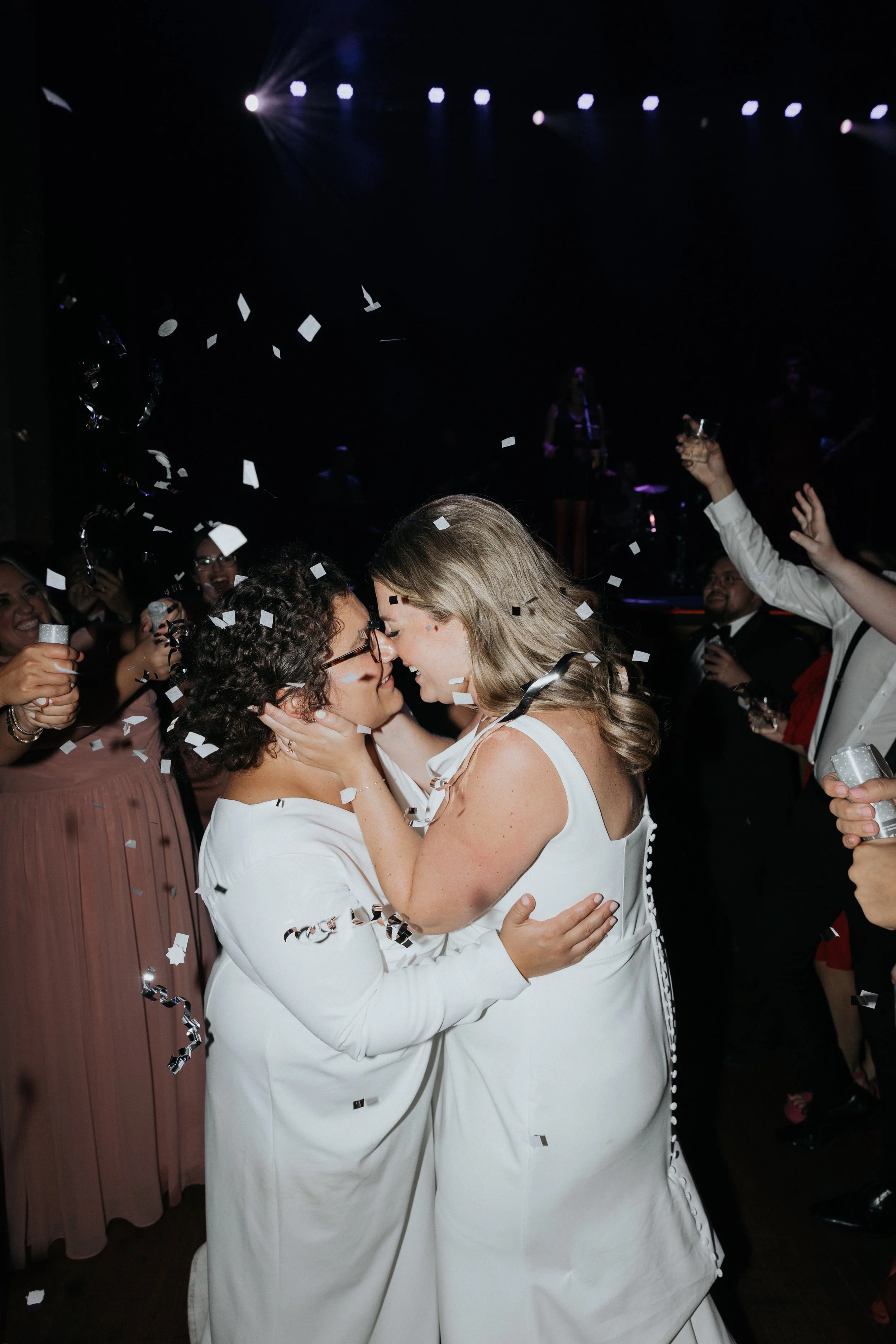 Two women in white dresses embrace each other in a dance at a celebration, surrounded by confetti with a dark stage and musicians in the background.