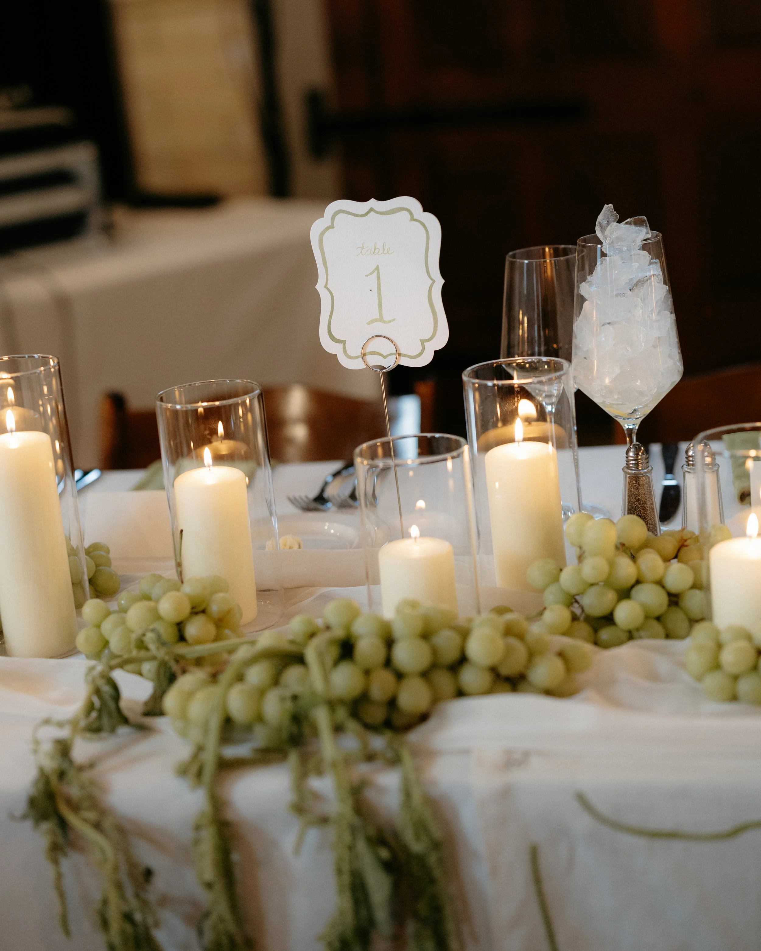 A wedding or event table setting with candles in glass holders, a framed table number card labeled 'Table 1', and a glass filled with ice. The table is decorated with white grapes and greenery.