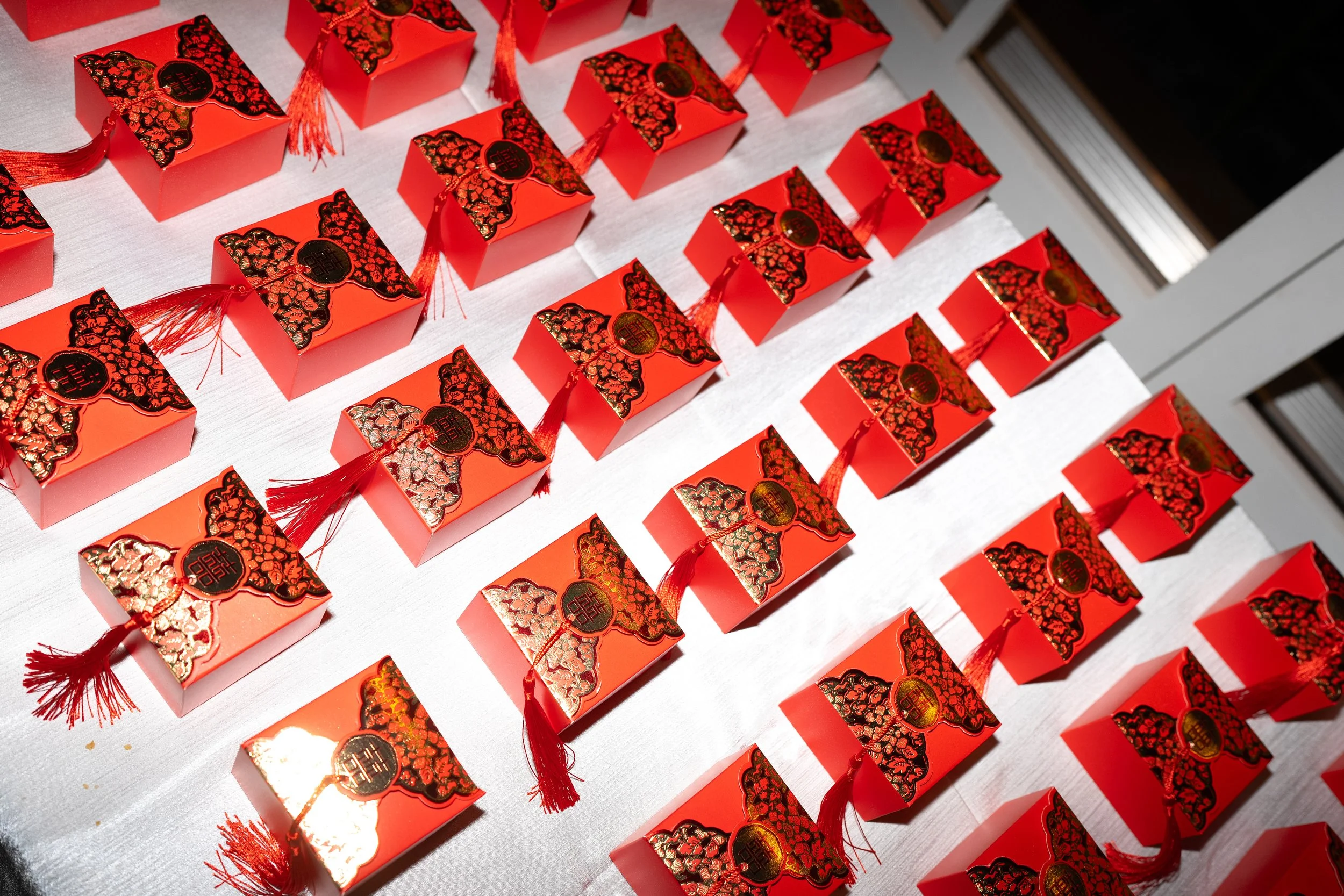 Rows of red Chinese gift boxes with gold and black decorative patterns, each tied with a red tassel, arranged on a table for a celebration or holiday.