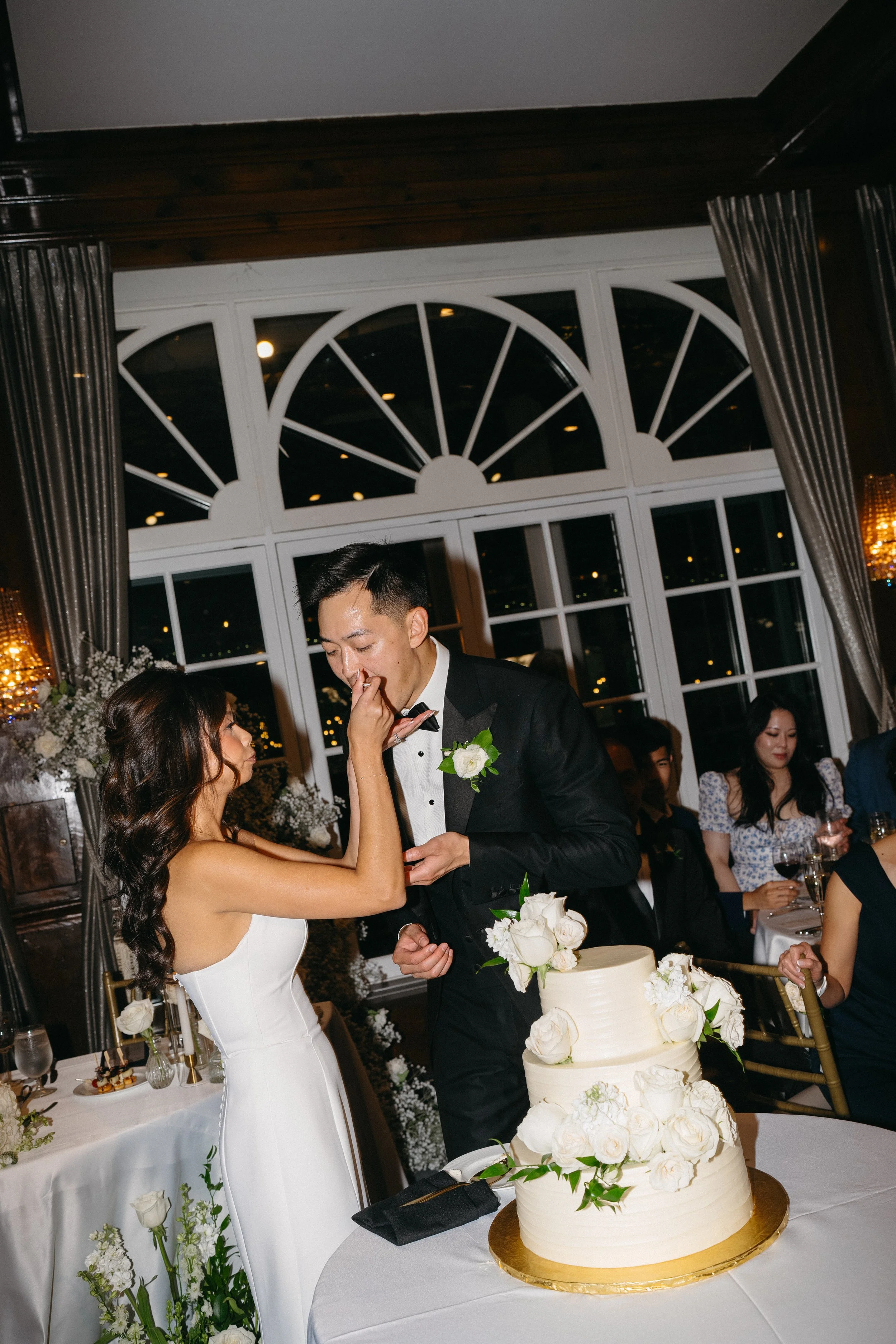 A bride and groom are cutting a wedding cake together at their reception. The bride is wearing a white strapless dress, and the groom is in a black tuxedo. There are guests seated at tables in the background, and the setting is decorated with flowers