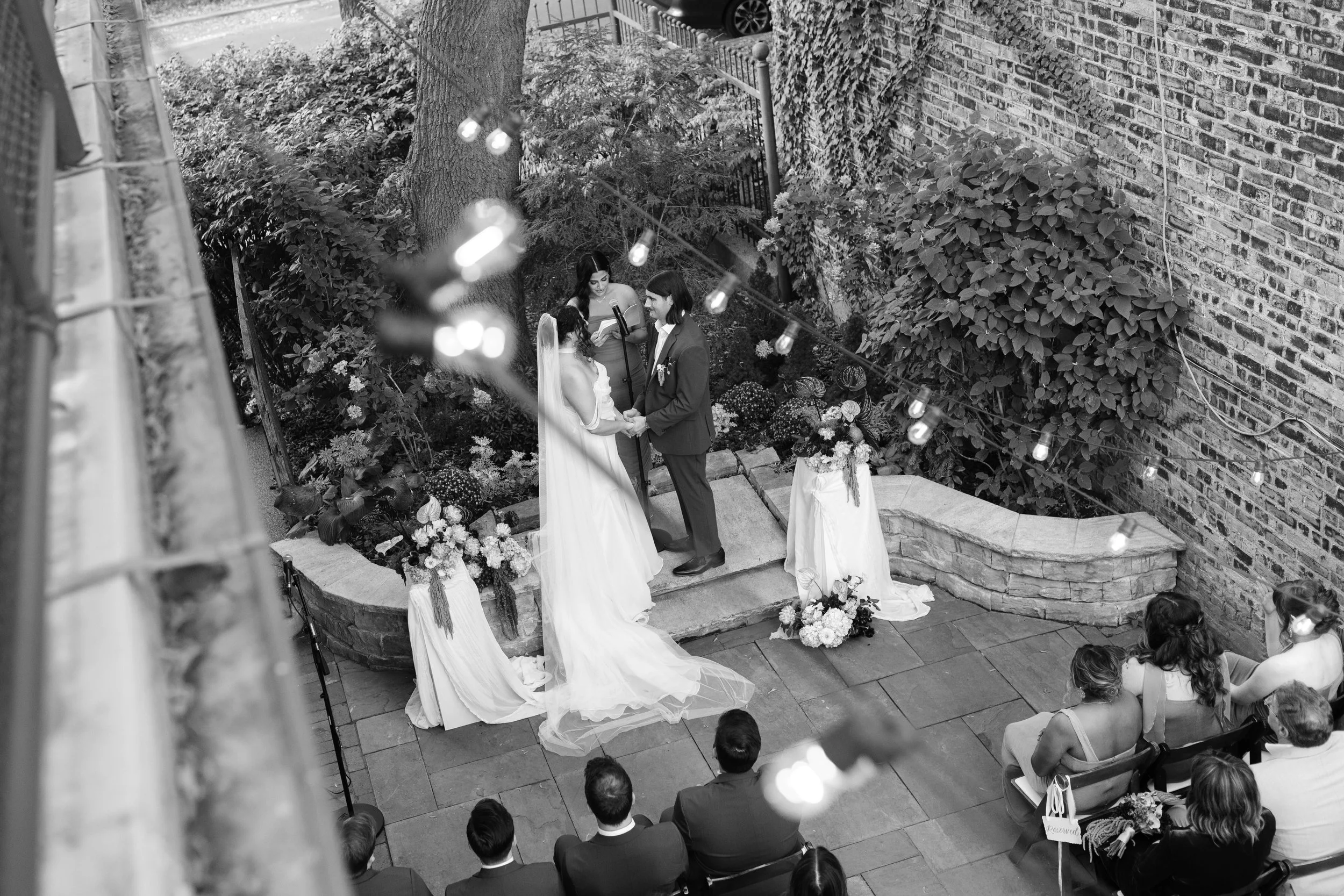 A black and white photo of a wedding ceremony taking place outdoors on a small stone patio with a bride and groom holding hands and facing each other, surrounded by guests seated in chairs. The ceremony is near a brick wall and underneath string ligh