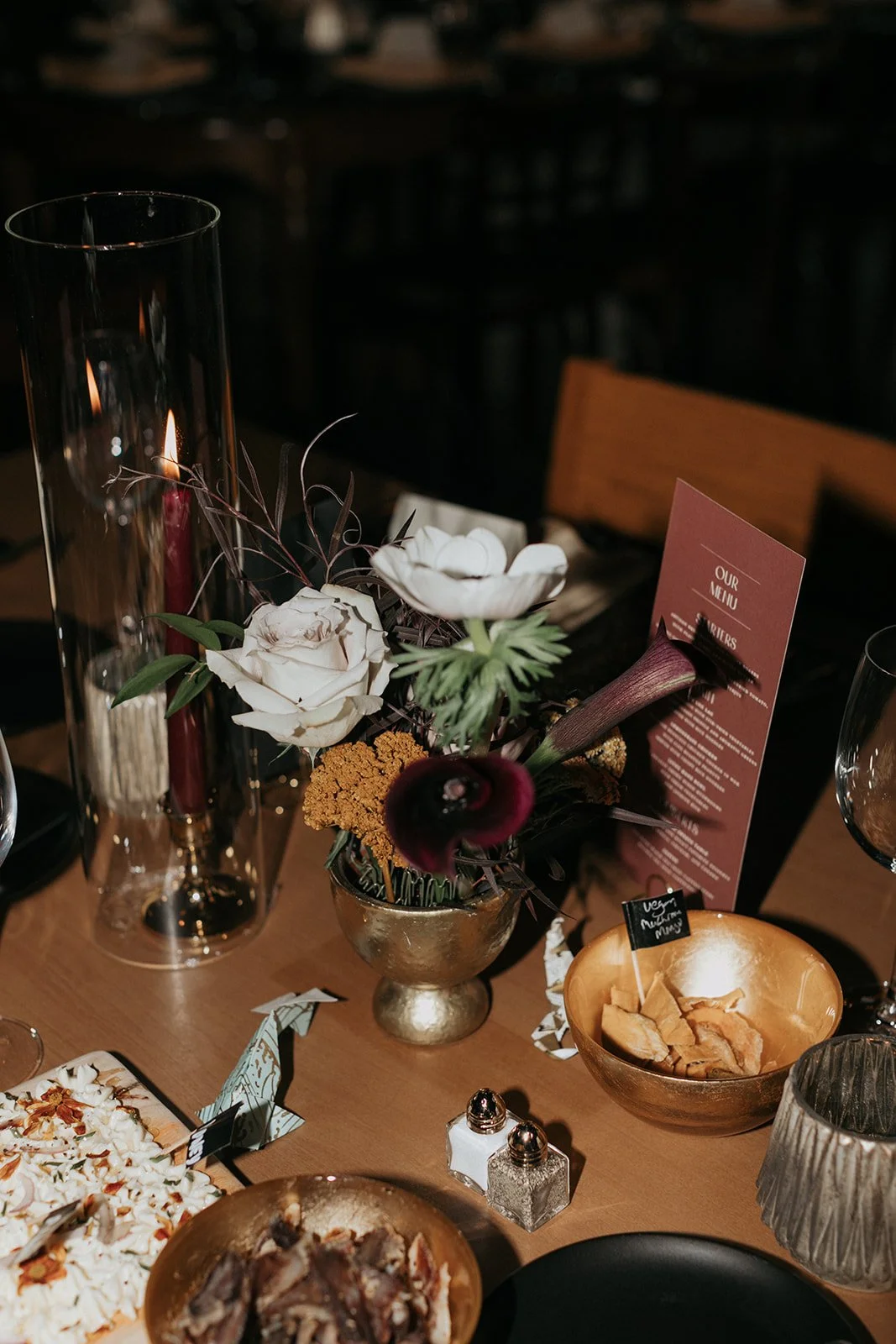 A table setting with a floral centerpiece, a candle in a tall glass holder, a menu card, and various dishes including pizza and chips at a dinner event.