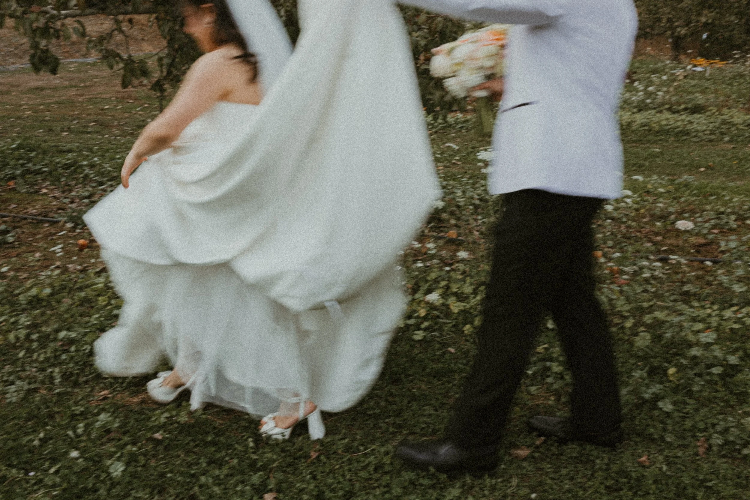 A couple dressed in wedding attire walking outdoors on grass with scattered flowers, one wearing a white wedding dress and the other in a light-colored outfit holding a bouquet.
