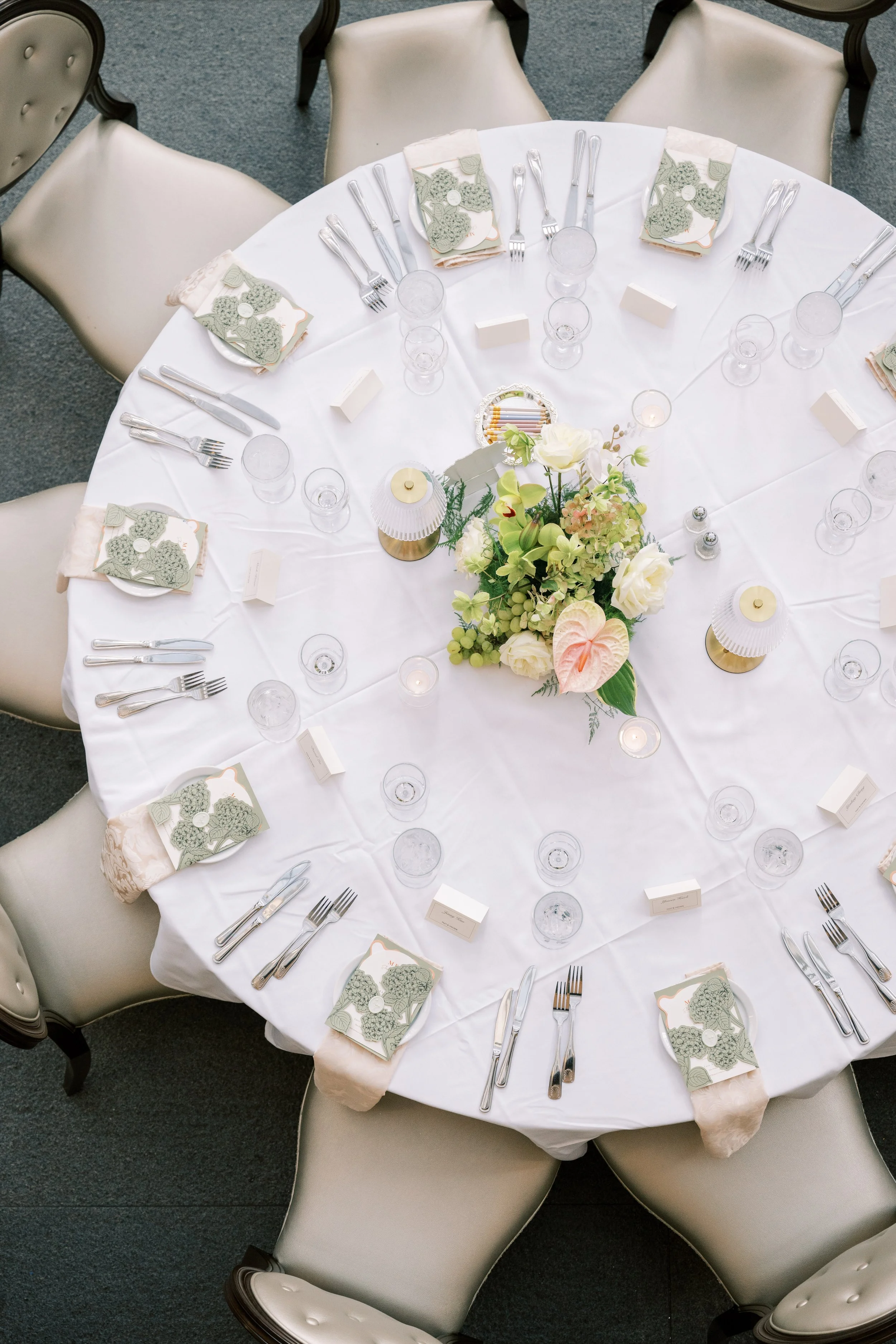 Round banquet table set for a formal event with white tablecloth, elegant chairs, floral centerpiece, place settings with forks, knives, spoons, glasses, and folded napkins decorated with green doilies.