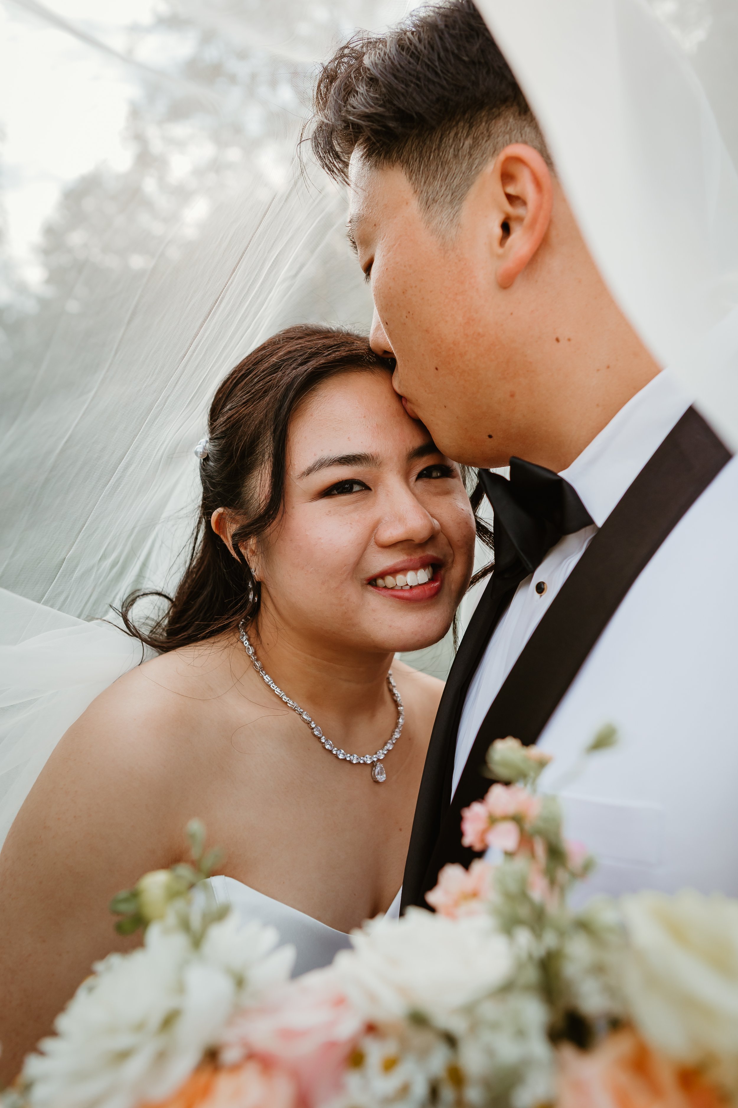 A bride and groom close-up portrait at wedding. The groom kisses the bride on forehead, she smiles with visible happiness. She wears a diamond necklace, the groom wears a tuxedo with a bow tie. Blurred floral bouquet is in foreground, background show