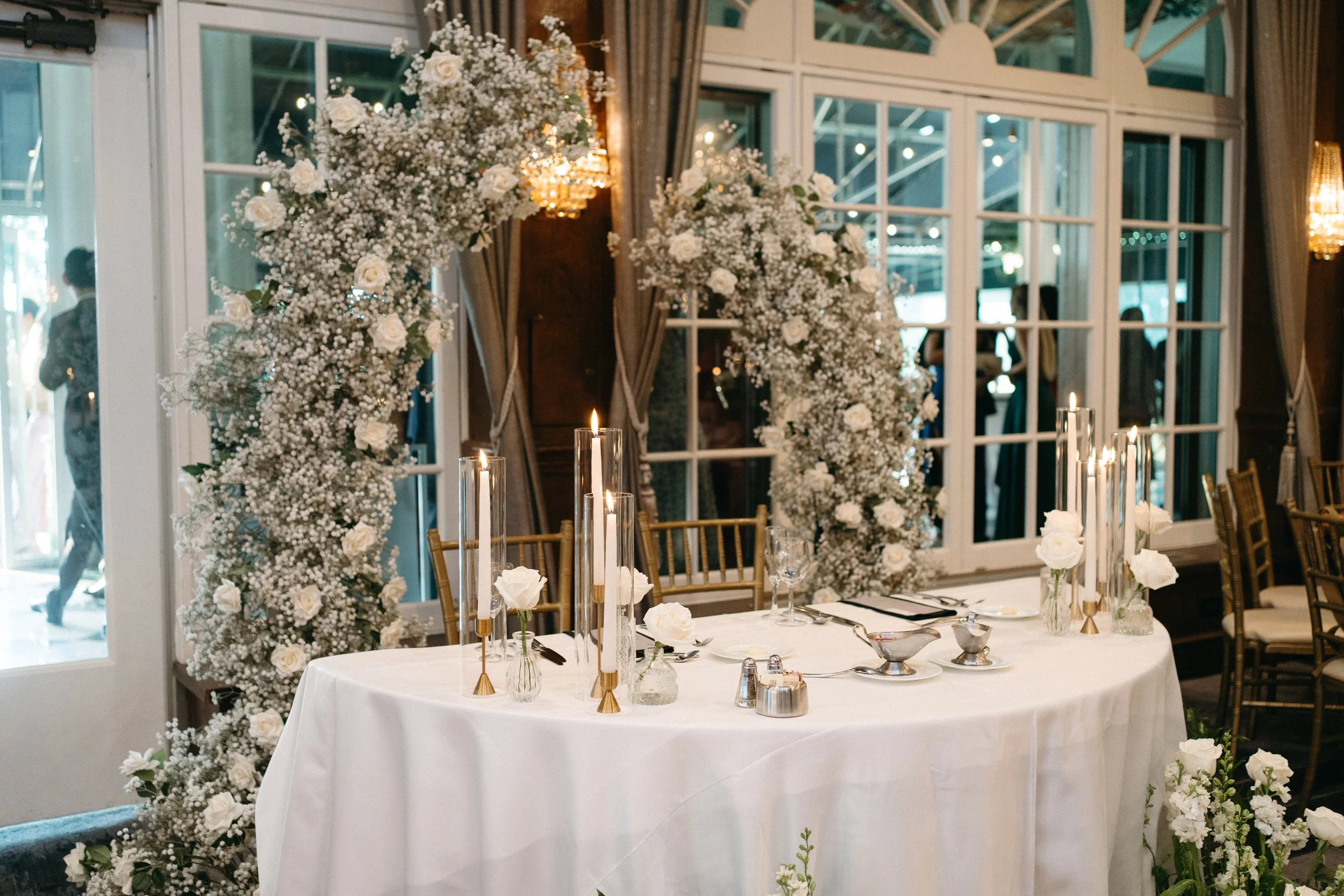 Elegant wedding reception setup with a white tablecloth, tall candles, white roses, and floral arrangements against large windows with draped curtains.