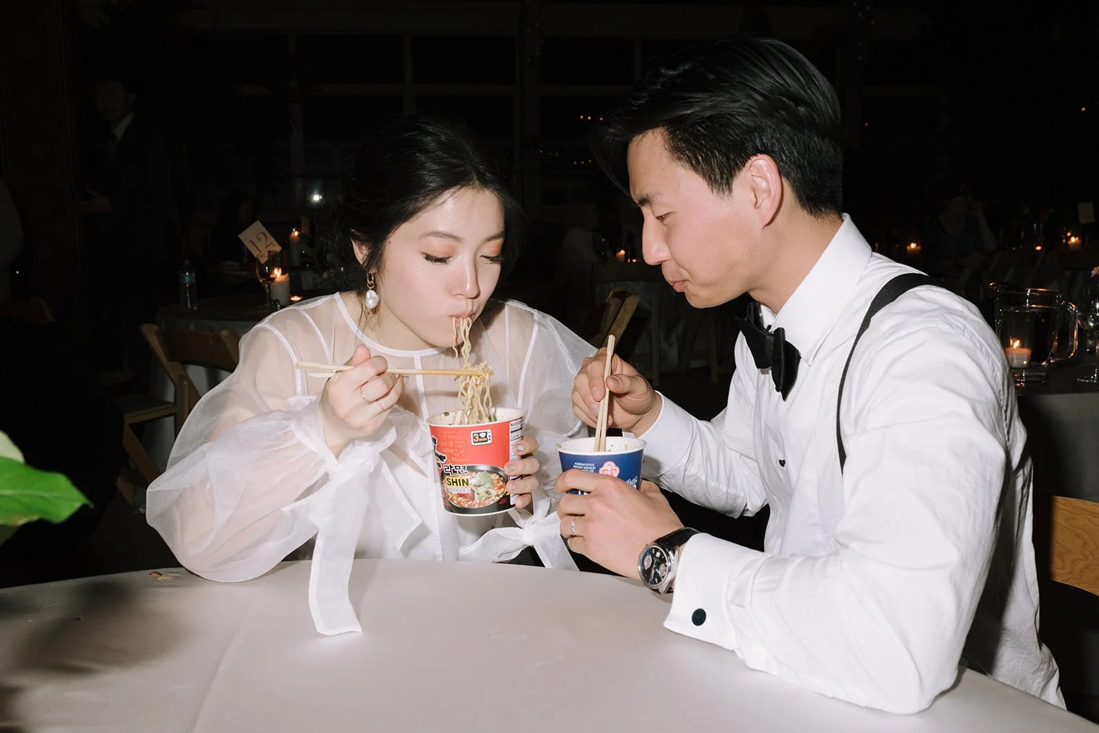 A man and woman in formal attire eating noodles from cups at a restaurant.