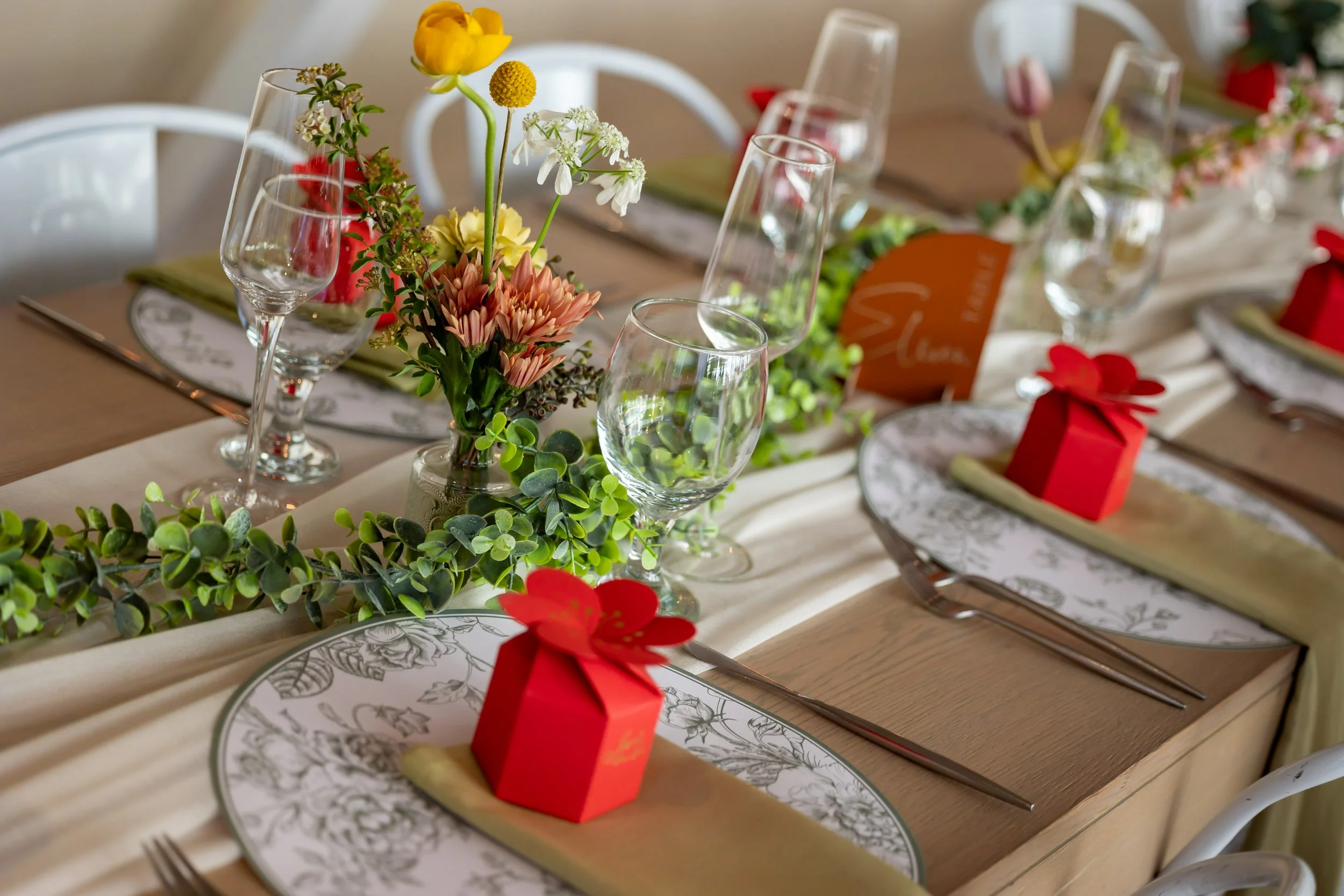 Table setting with floral centerpiece, wine glasses, paper plates with floral design, greenery garland, and red gift boxes with a bow, set on a wooden table.