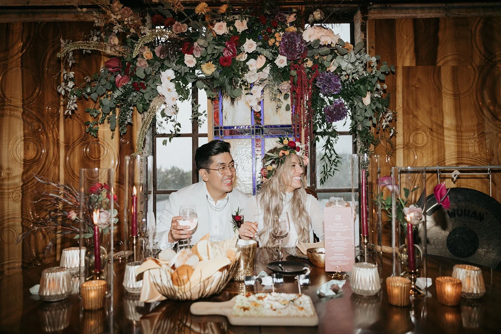 A couple sitting at a decorated dining table with candles, a floral arrangement, and a platter of food, celebrating under a floral canopy, smiling and holding drinks.