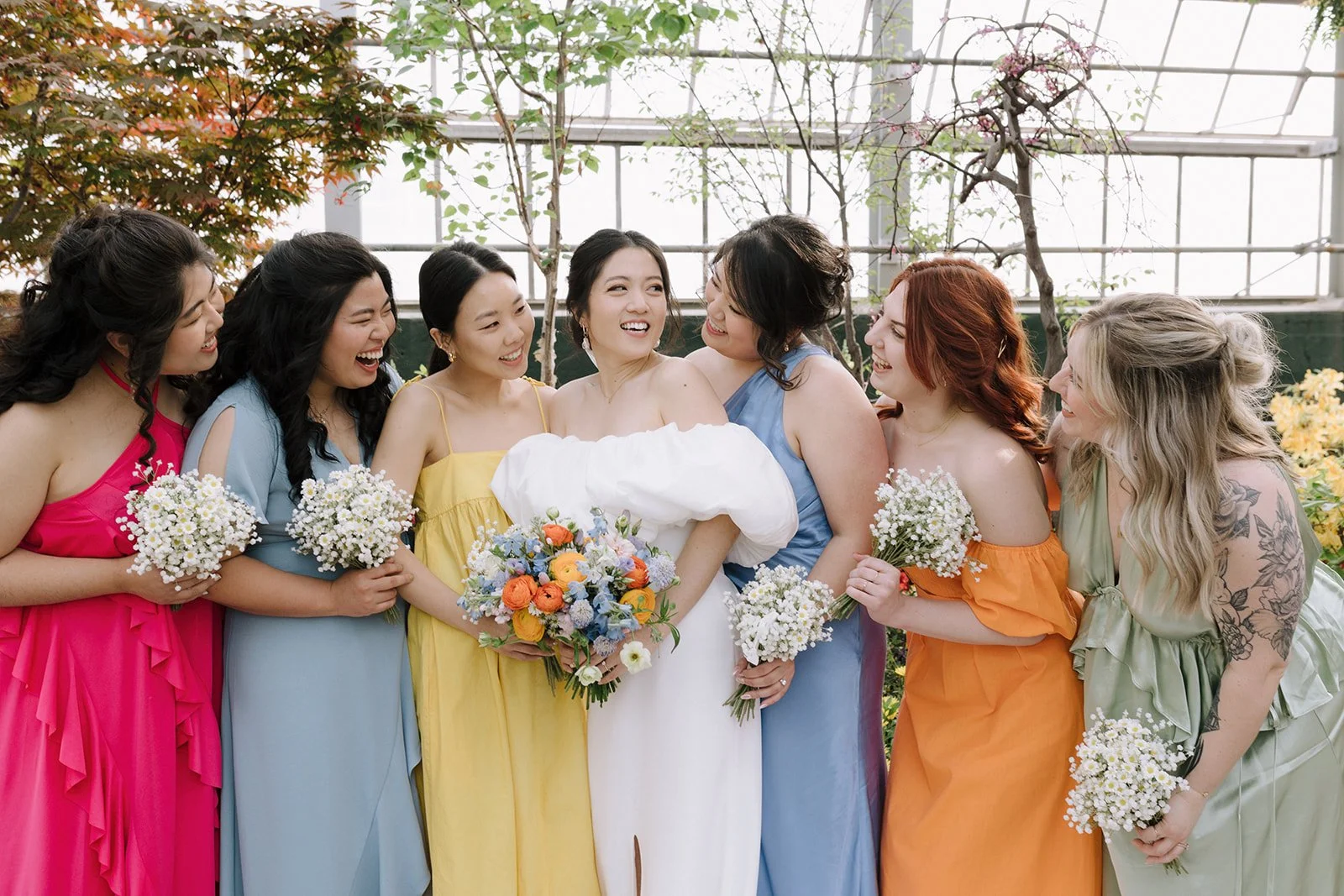 A bride surrounded by seven women in colorful dresses holding flowers, inside a greenhouse.