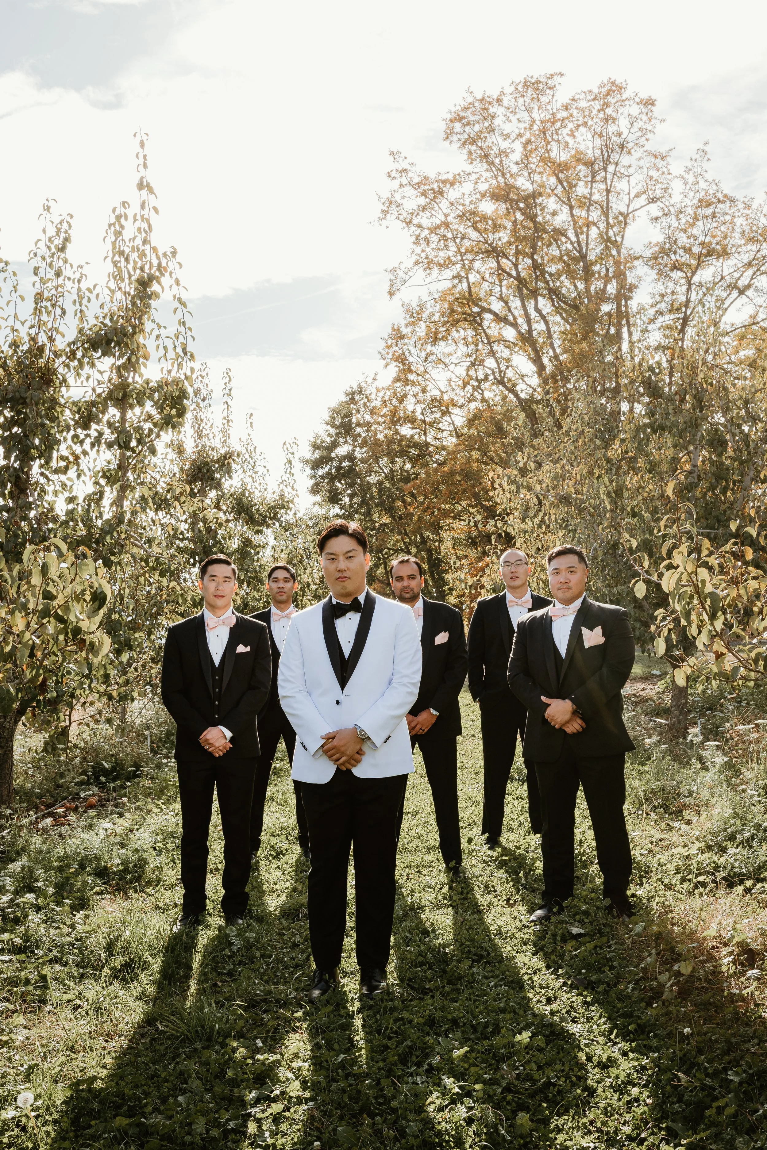A group of six men in tuxedos walking outdoors through grass and trees, with one man in a white tuxedo in the center and the others in black tuxedos with light pink bow ties, during daytime.