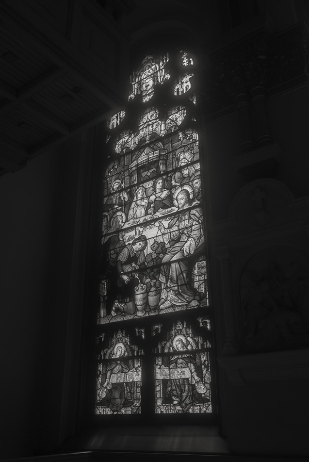 Black and white photo of a stained glass window depicting a religious scene with multiple figures, some with halos, inside a church.