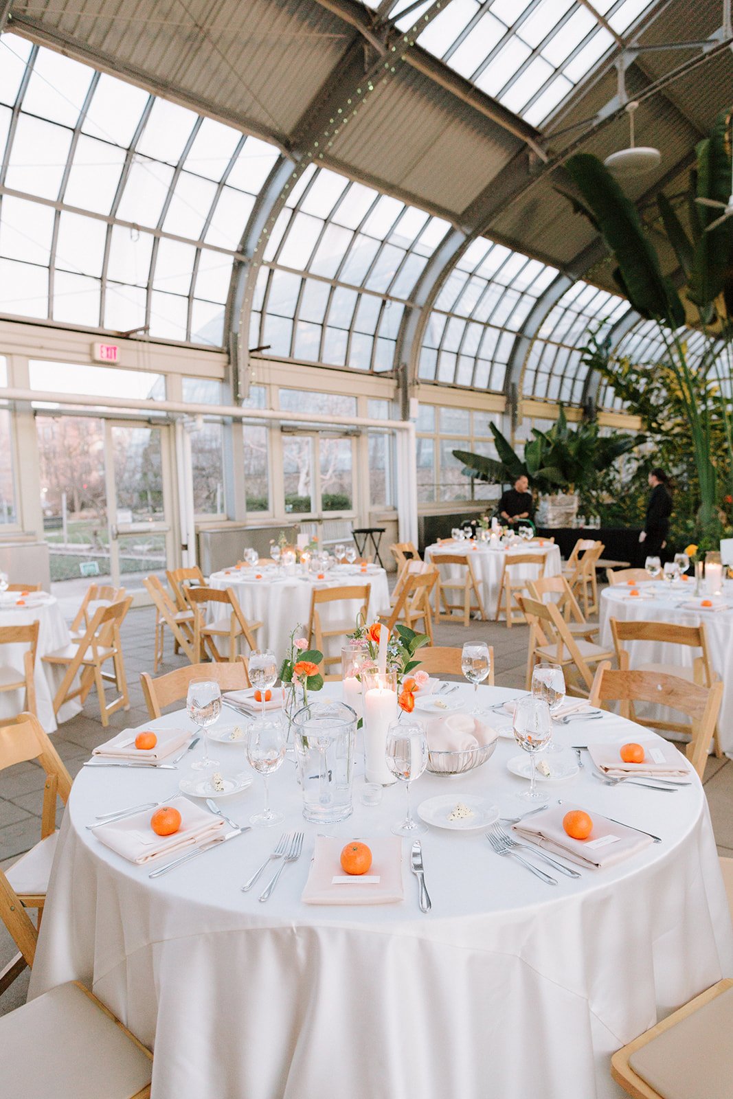 Elegant indoor event space with round tables draped in white tablecloths, set with silverware, water glasses, small orange fruits, and floral centerpieces. The space features large glass windows and greenery.