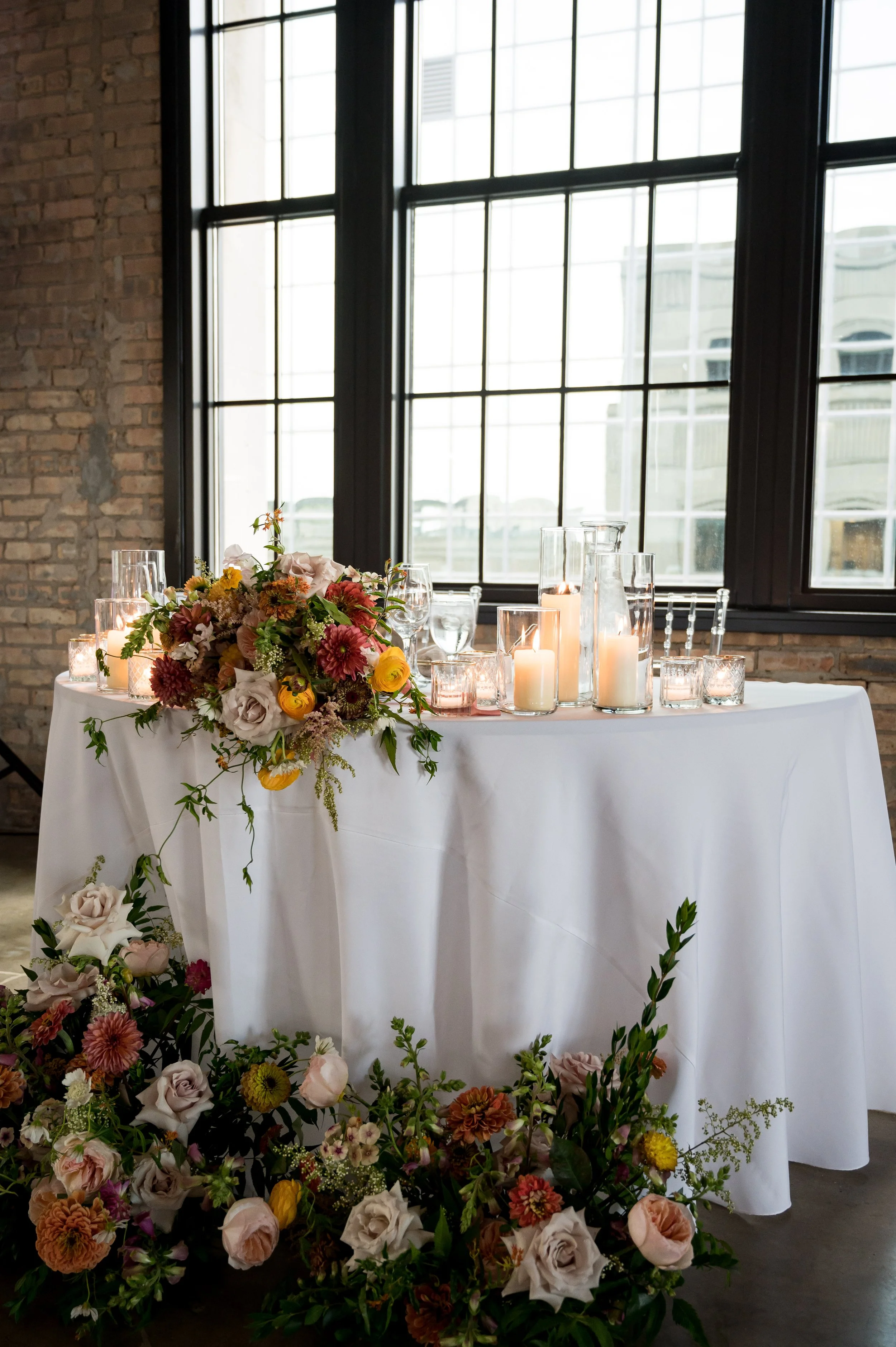 A table decorated with a large floral arrangement and candles in glass holders, set against a brick wall and large multi-pane windows.