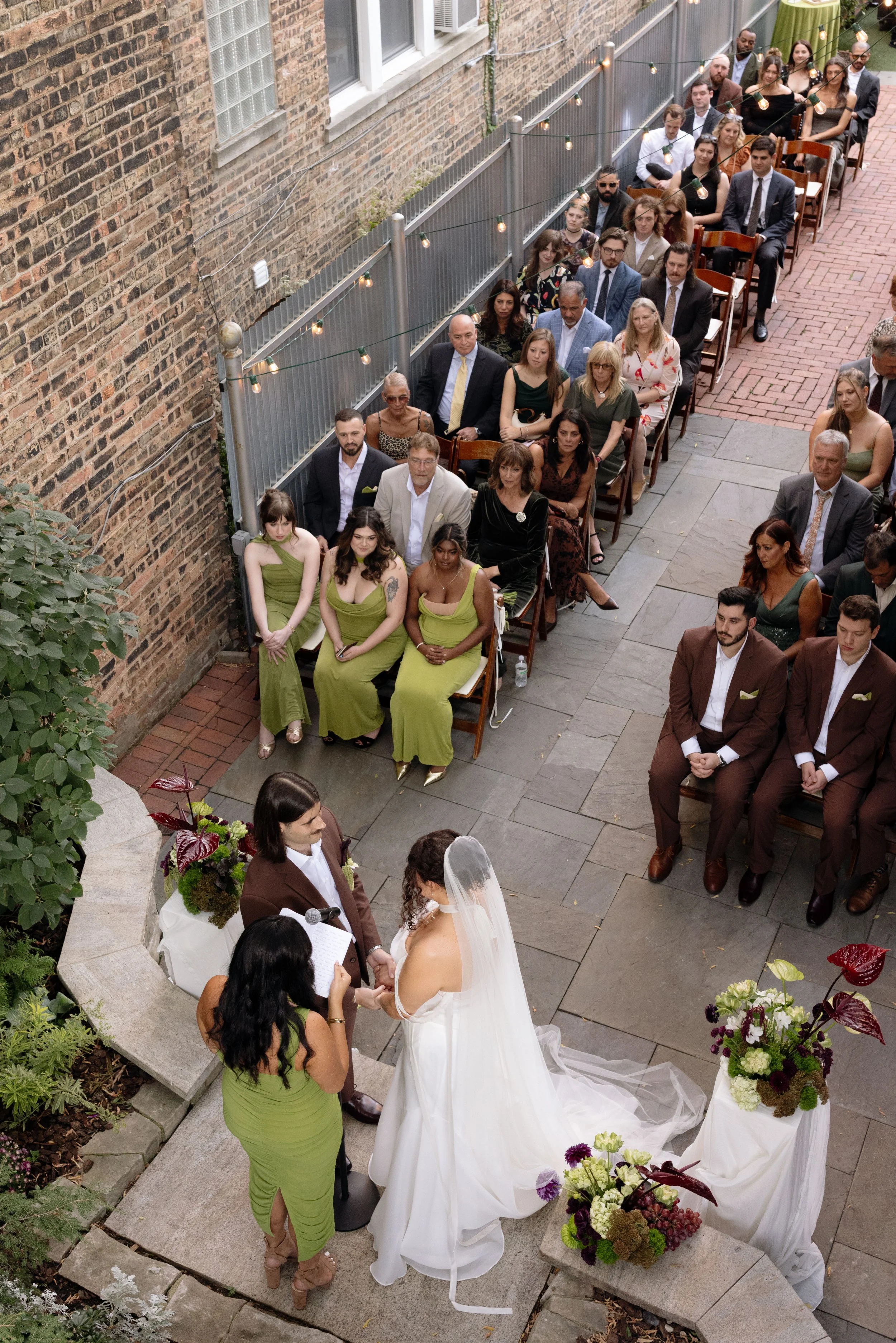 Wedding ceremony outdoors with bride and groom holding hands, officiant, and guests seated in rows. Decor includes flowers and string lights alongside a brick wall.