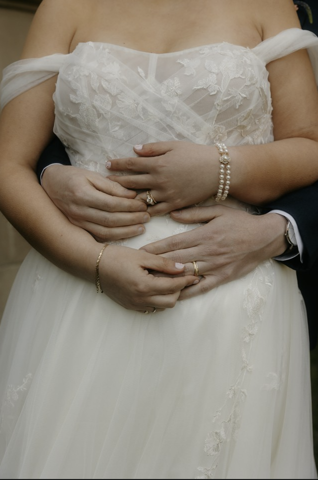 Close-up of a bride and groom's hands forming a heart shape on her wedding dress, with the bride's cathedral-length lace gown and jewelry visible.
