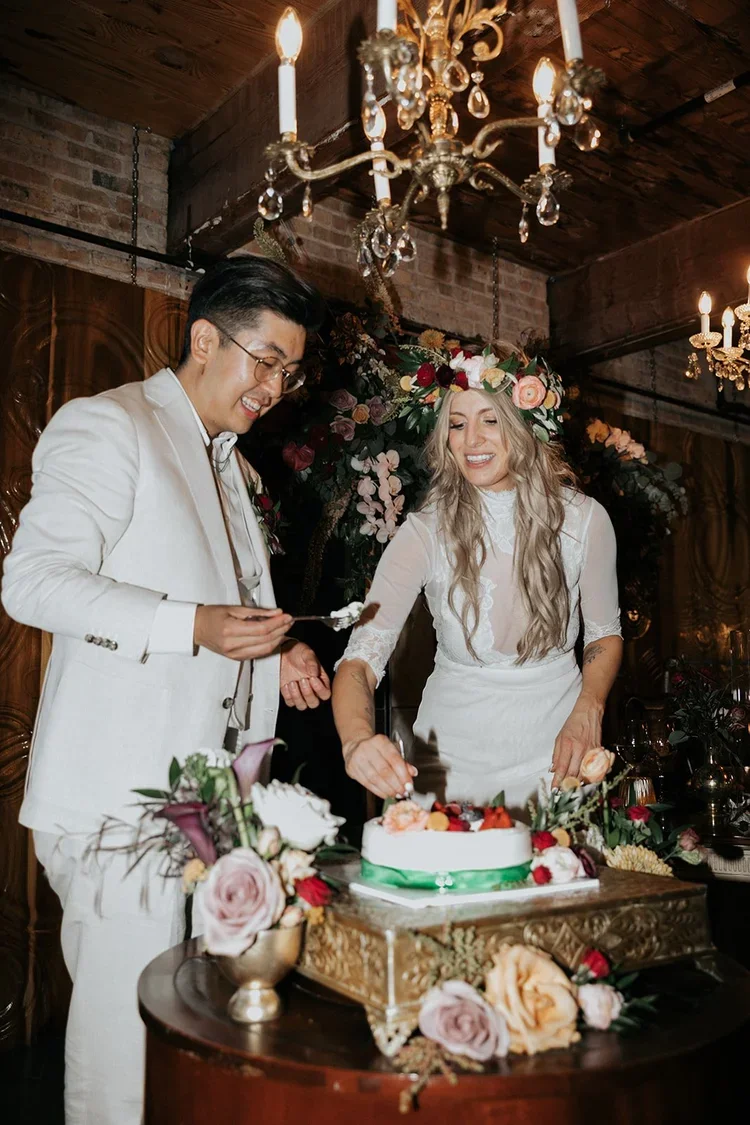 Couple at a wedding reception, with the bride wearing a flower crown and white dress, and the groom in a white suit, cutting a wedding cake decorated with fresh flowers, in an elegant, dimly lit room with chandeliers and floral arrangements.
