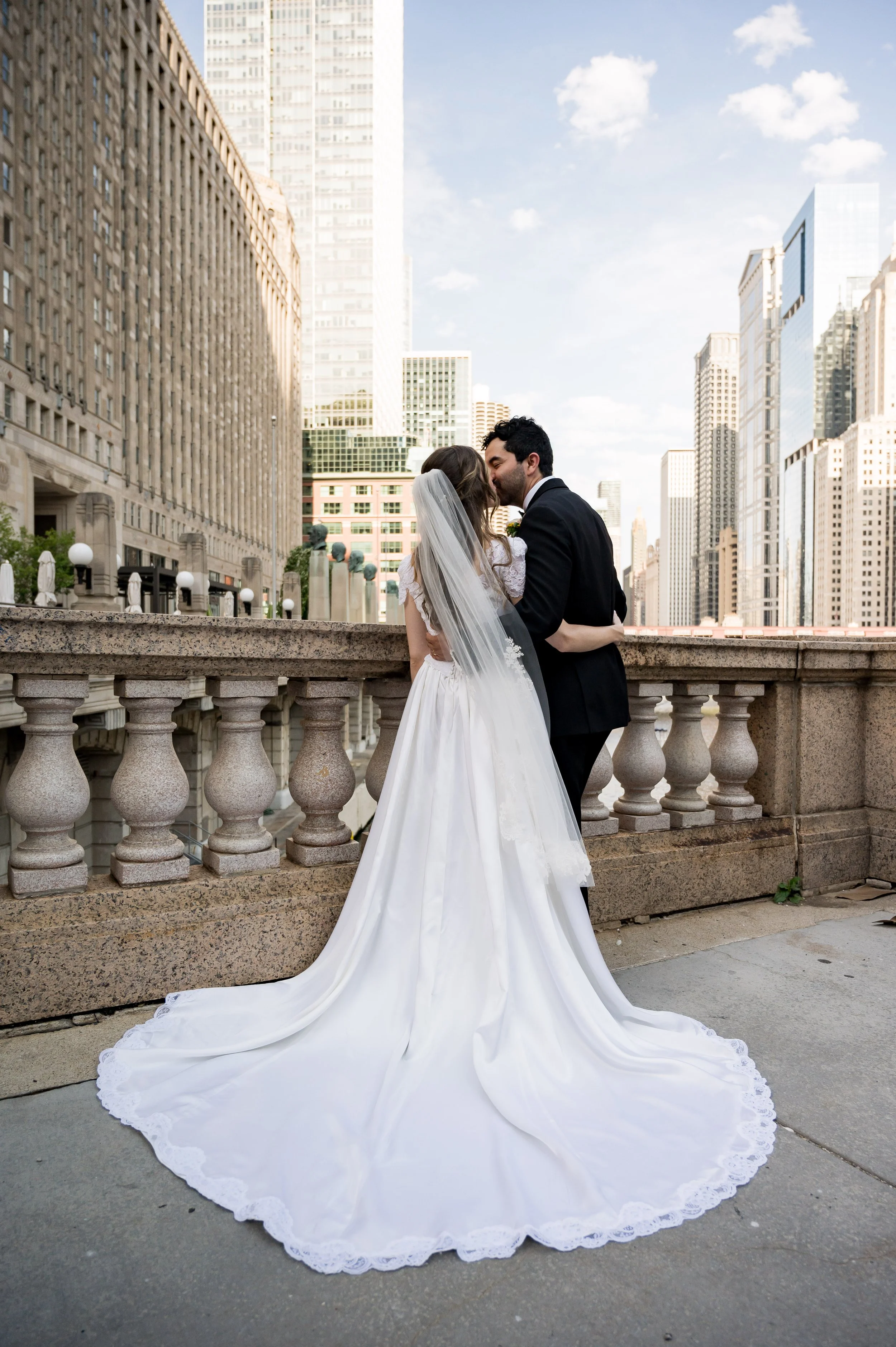A newlywed couple sharing a kiss on a city bridge, with the bride wearing a long white gown with lace details and a veil, and the groom in a black suit, against a backdrop of tall skyscrapers and a blue sky with some clouds.