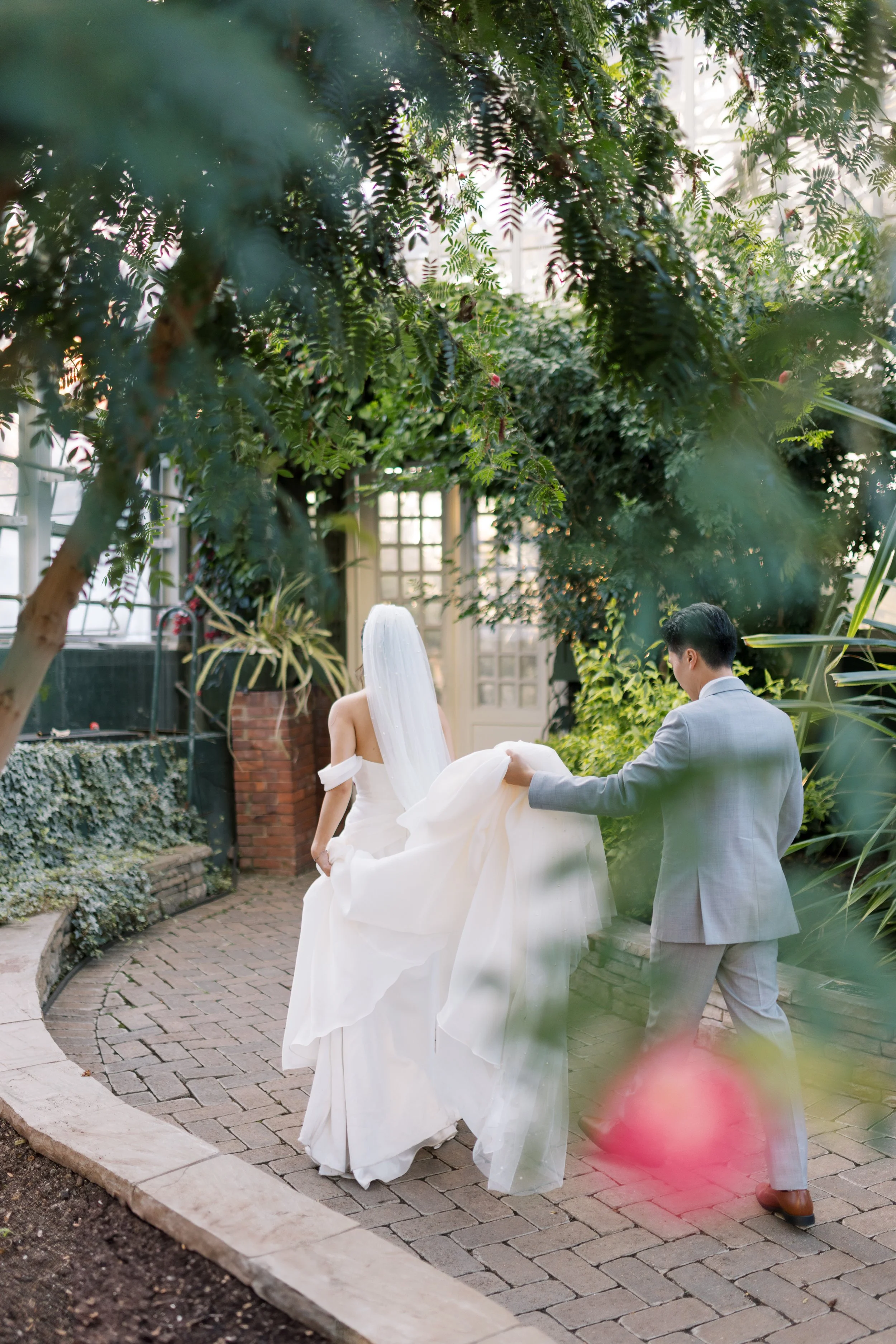 A bride and groom holding hands and dancing outdoors in a garden-like setting with lush green trees and brick paving, with a white door in the background.