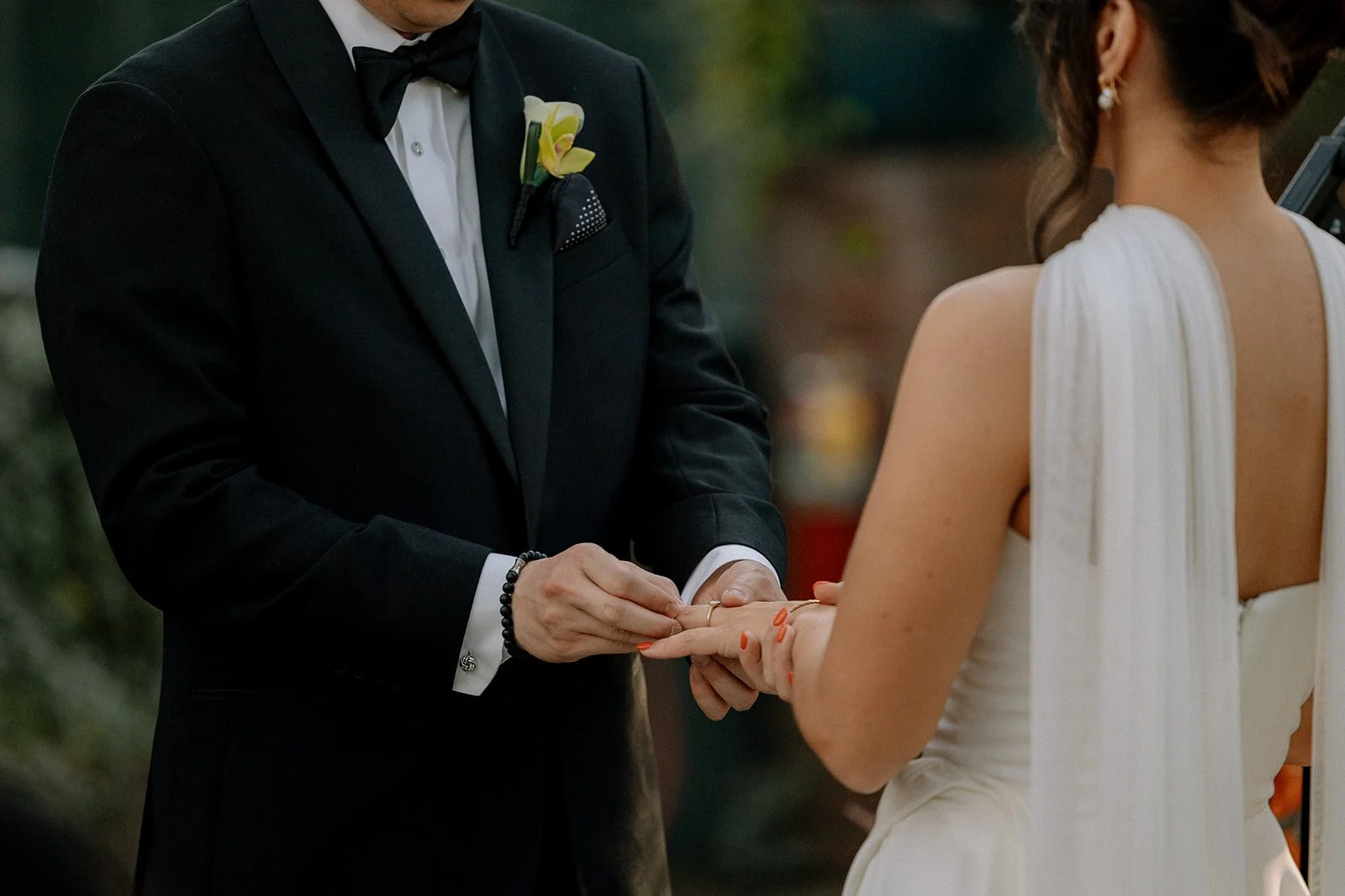 A couple is exchanging wedding vows outdoors; the groom is placing a ring on the bride's finger. The groom is dressed in a black tuxedo with a black bowtie, and the bride wears a white wedding gown with a pleated back. The scene is likely part of a w
