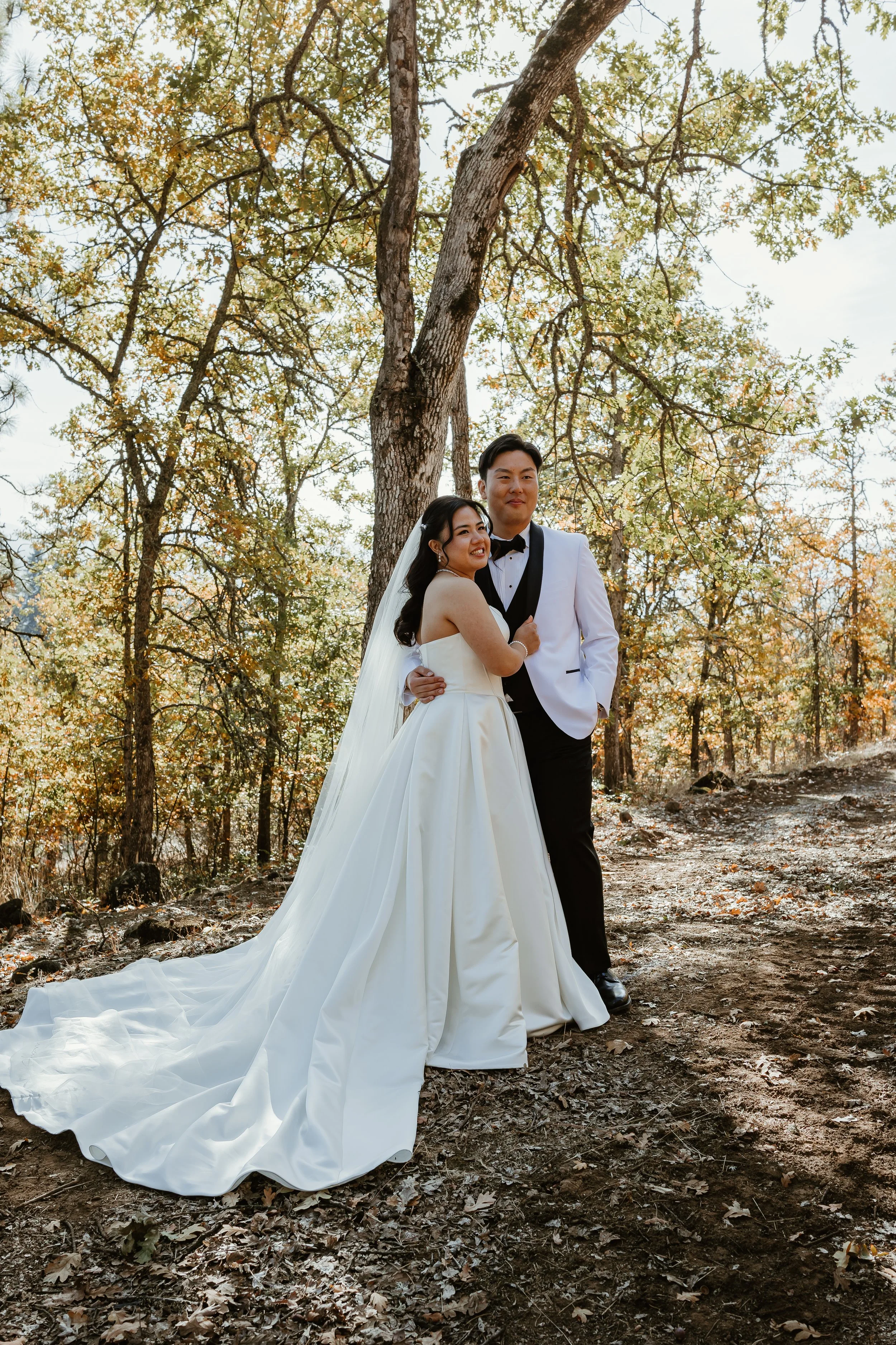 A newlywed couple smiling and posing in a forest during autumn, with the bride in a white wedding gown and veil, and the groom in a white tuxedo jacket with black lapels and black pants, standing close together.