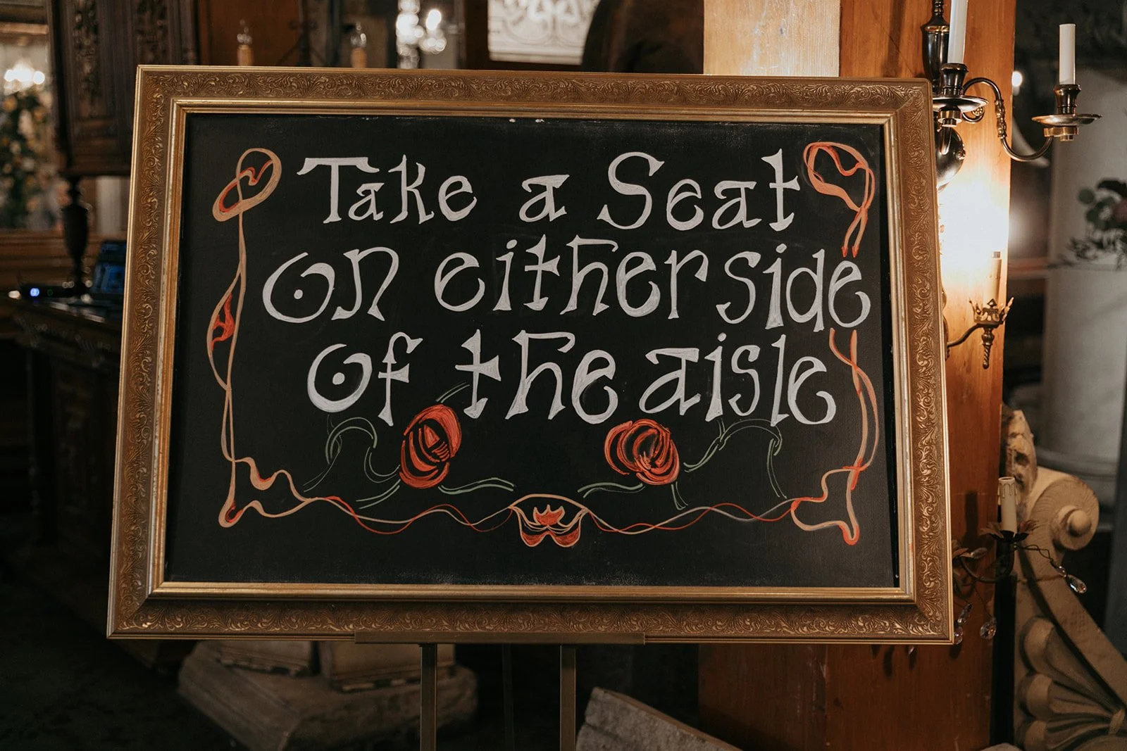 A decorative black chalkboard with a gold ornate border, featuring the handwritten message: "Take a Seat on either side of the aisle" in white and orange chalk. The board is placed on a stand and is illuminated by candle-style wall sconces on the rig