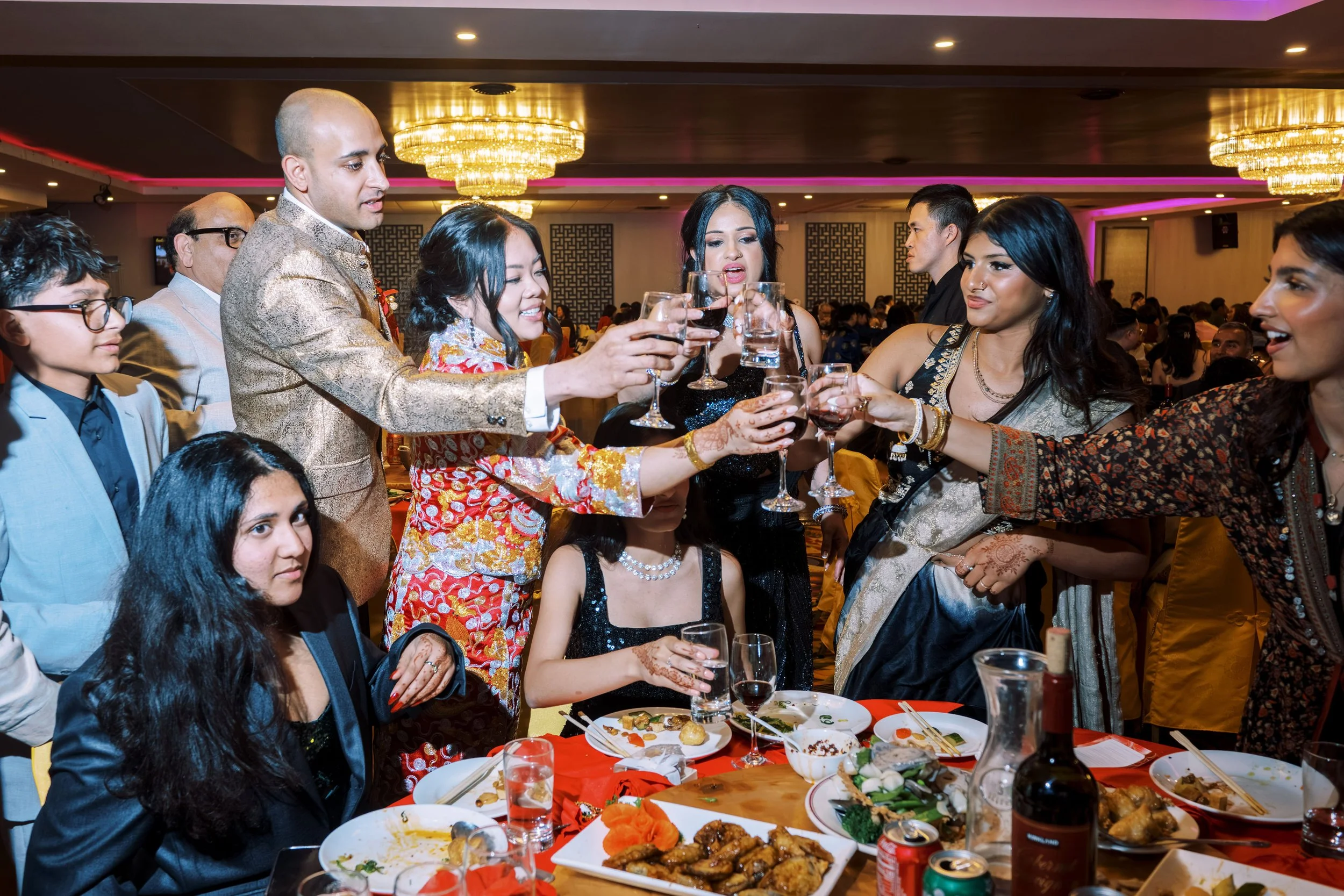 A group of people celebrating at a banquet, toasting with wine glasses, with food on the table, in a decorated banquet hall with chandeliers.
