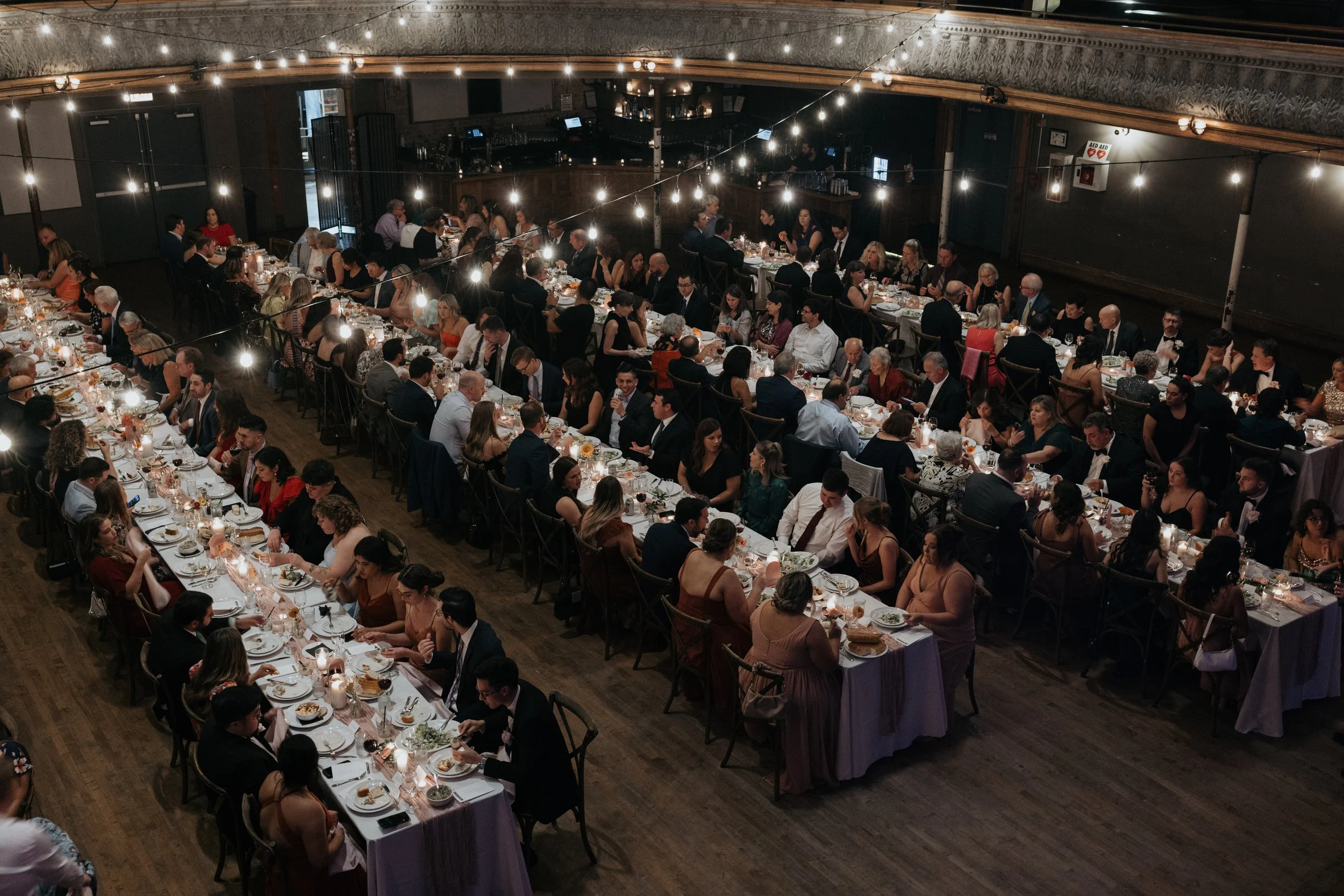 A large banquet hall filled with long tables where a diverse group of people are dining, under string lights hanging from the ceiling, with a bar area in the background.
