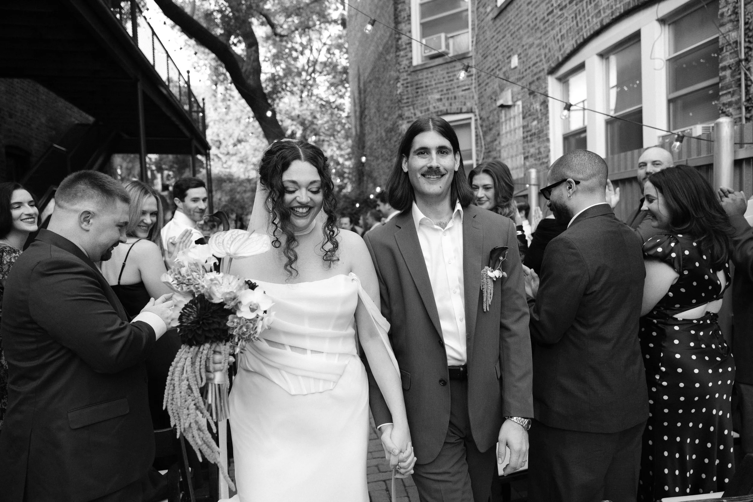 A black-and-white photo of a wedding ceremony outdoors, with a bride and groom holding hands and standing among friends and family. The bride is wearing a white dress and holding a bouquet, and the groom is in a suit. Guests are gathered around, smil