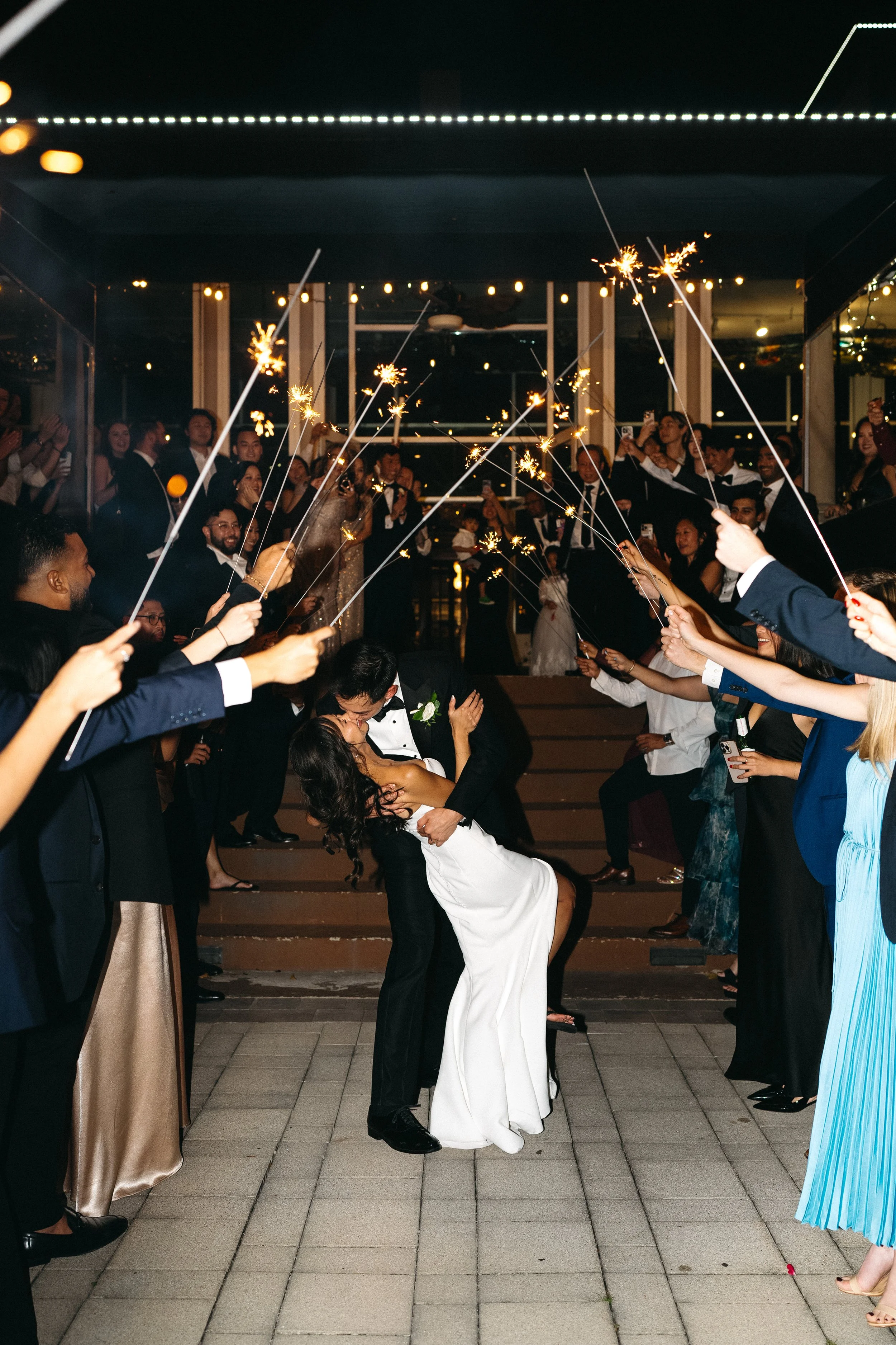 People in formal attire celebrating with sparklers at a wedding reception on stairs, with a woman in a white dress kneeling and a man in a tuxedo.