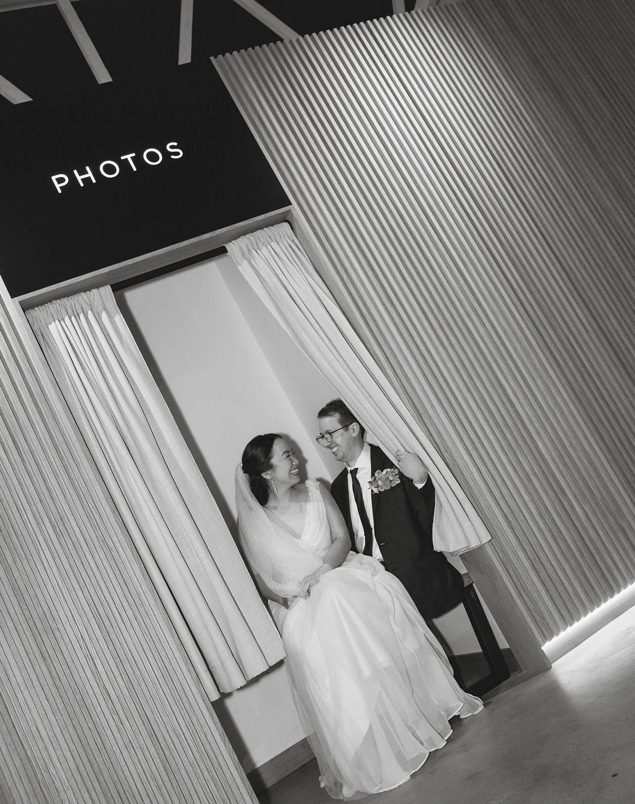 Black and white photo of a bride and groom sitting together on a stage framed by curtains, smiling at each other at their wedding reception. Behind them are vertical striped curtains and a sign that reads "PHOTOS".