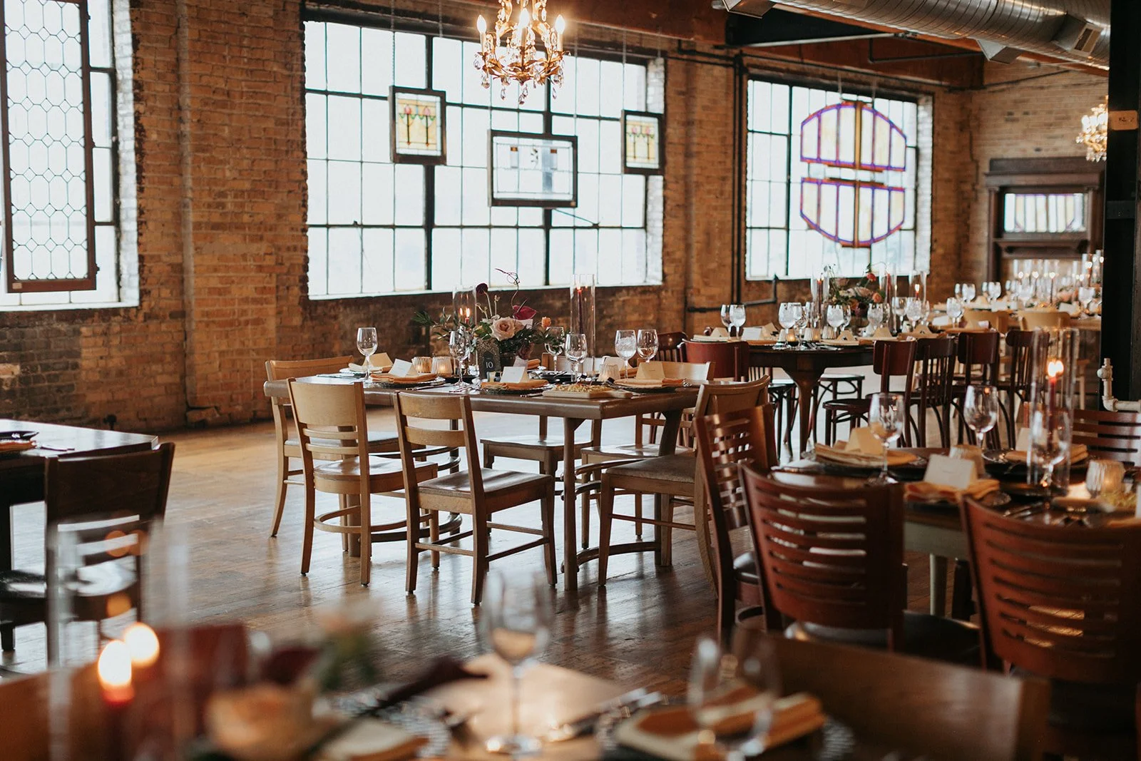 Elegant dining area with wooden tables and chairs, set for a formal event, inside a brick-walled venue with large stained glass windows and chandeliers hanging from the ceiling.