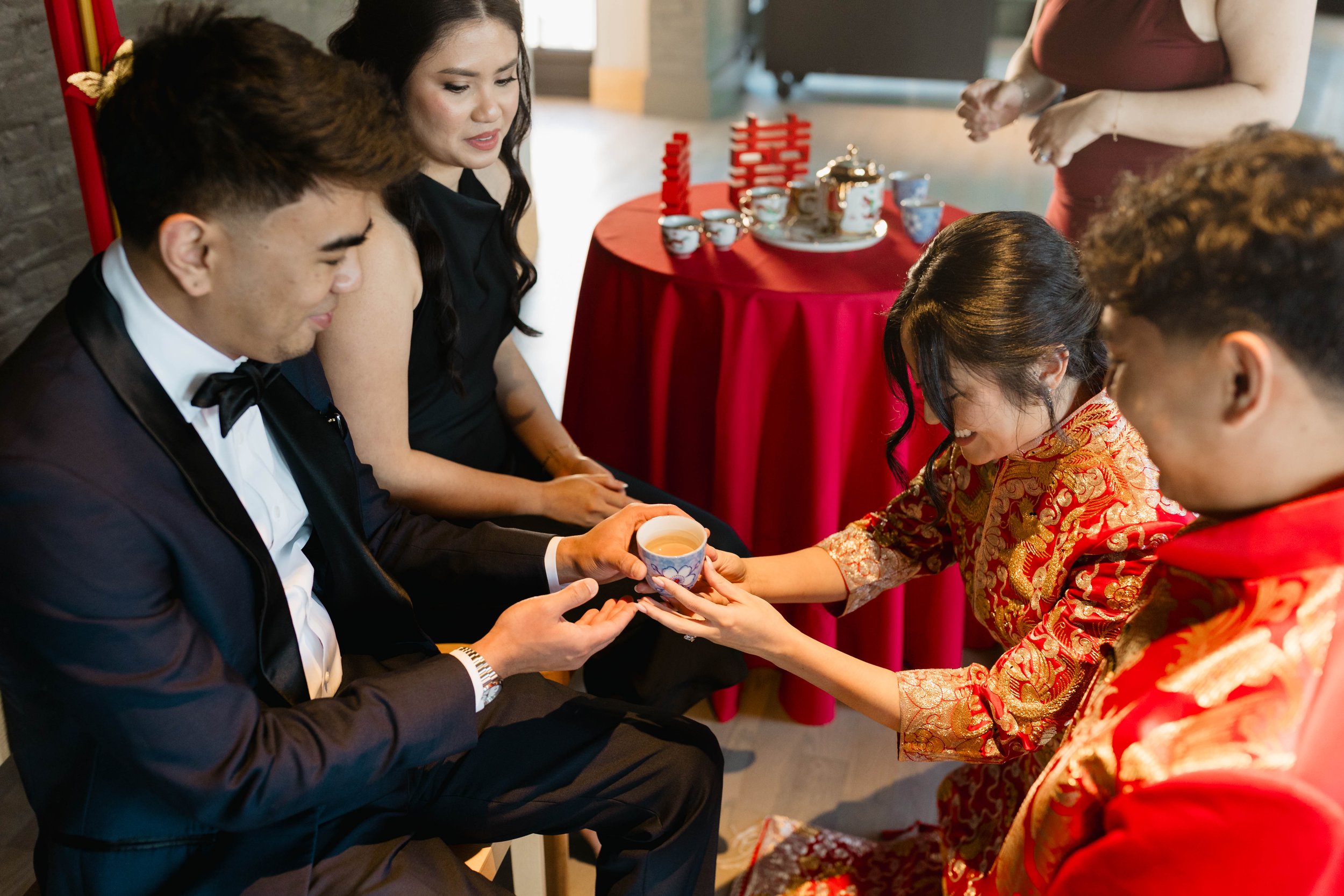 A traditional Chinese tea ceremony with a woman in a red and gold embroidered dress presenting tea to a man in a tuxedo, in a room decorated with red accents and teaware on a red table.