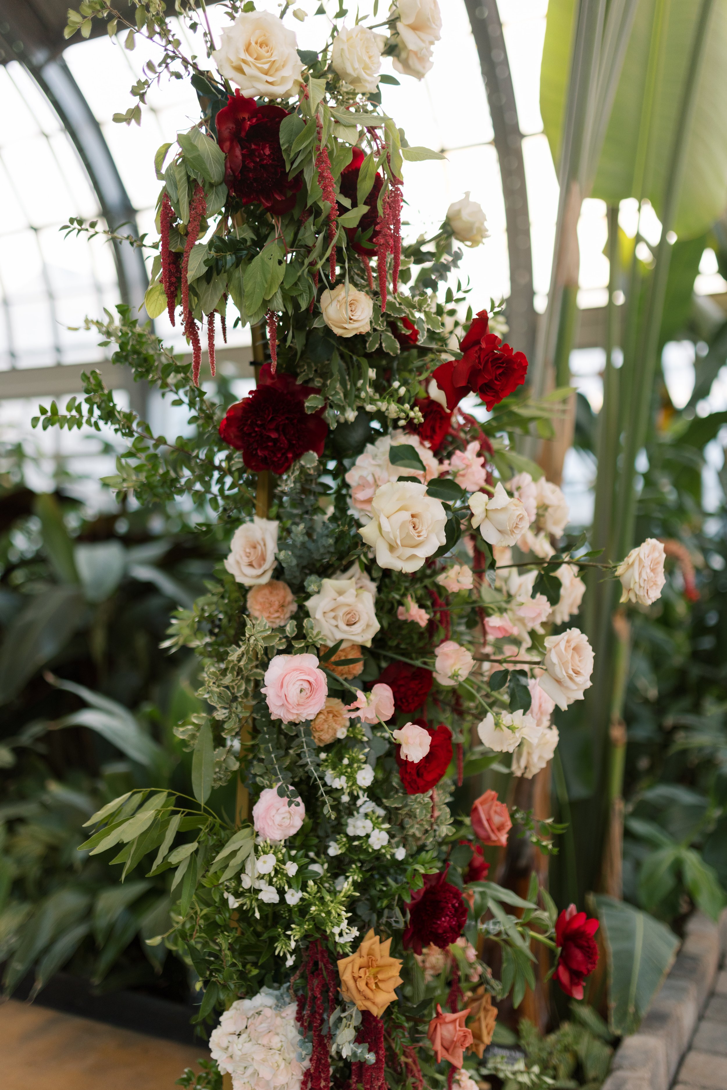 A vertical floral arrangement featuring white, red, pink, peach, and yellow flowers, including roses and carnations, with green foliage, set against a greenhouse background.