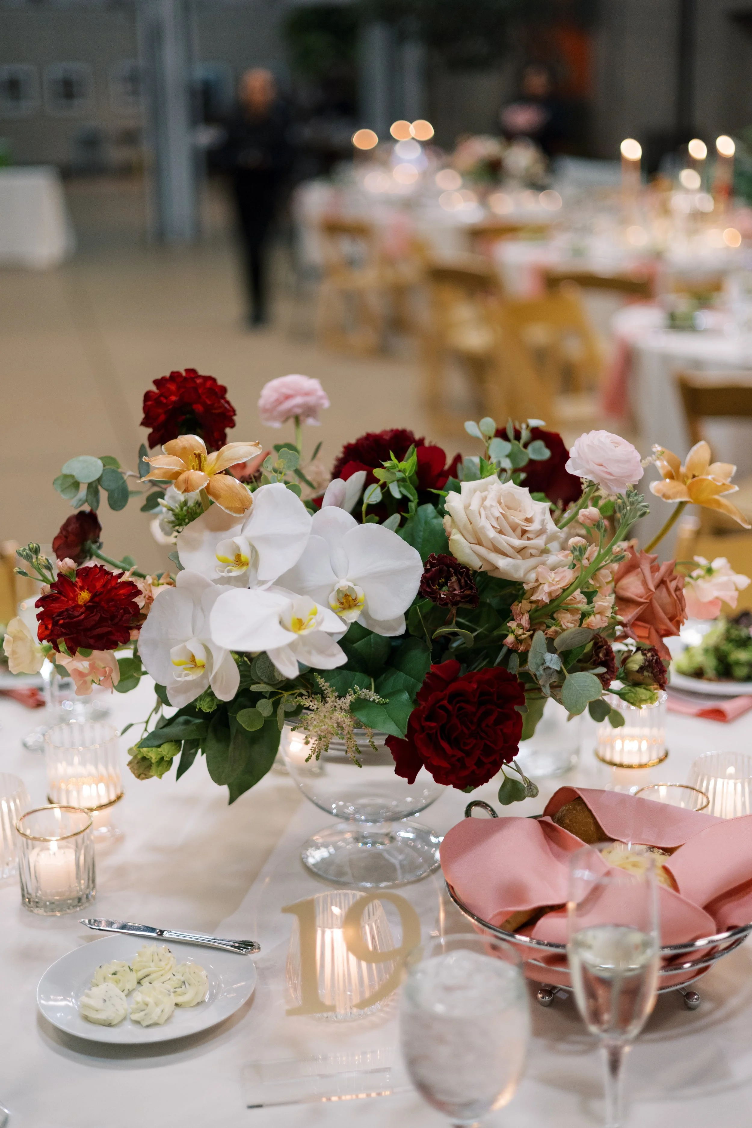 A close-up of a floral centerpiece with roses, orchids, and other flowers on a banquet table at a formal event with blurred background of chairs, tables, and people.