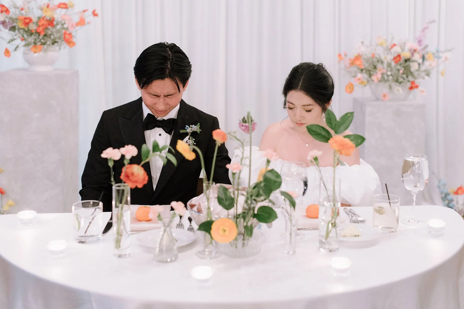 A bride and groom sit at a decorated wedding reception table with flowers and glasses, bowing their heads in prayer.